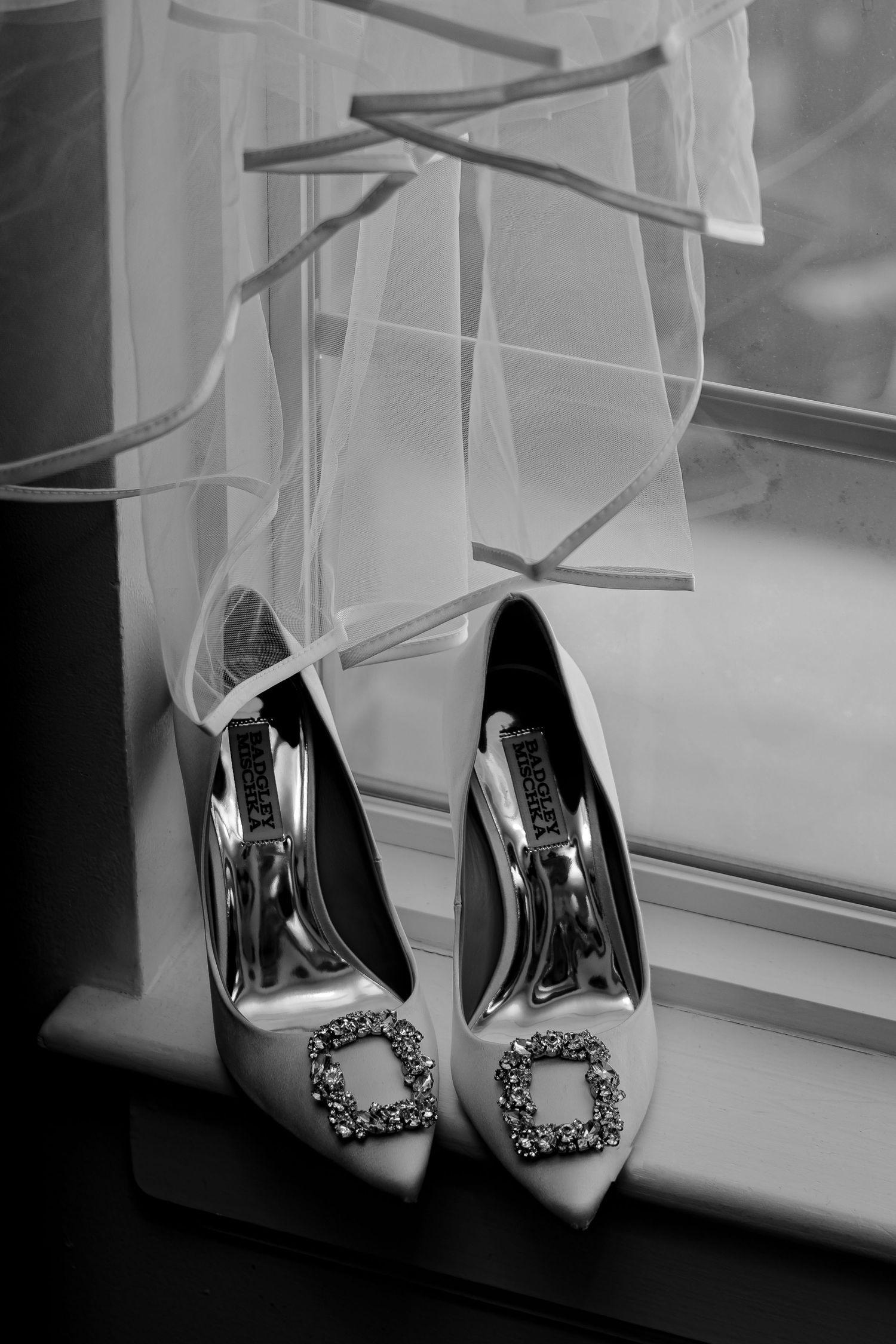 Black and white photo of bridal heels with crystal buckles on windowsill with wedding veil.