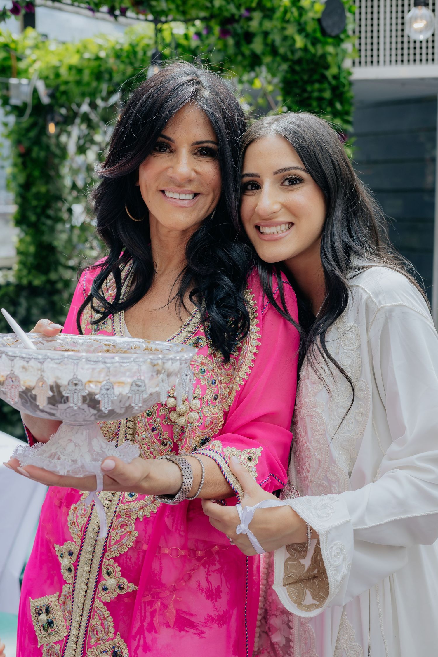 Two women, one in pink traditional attire and one in white, smile while holding a decorative crystal bowl.
