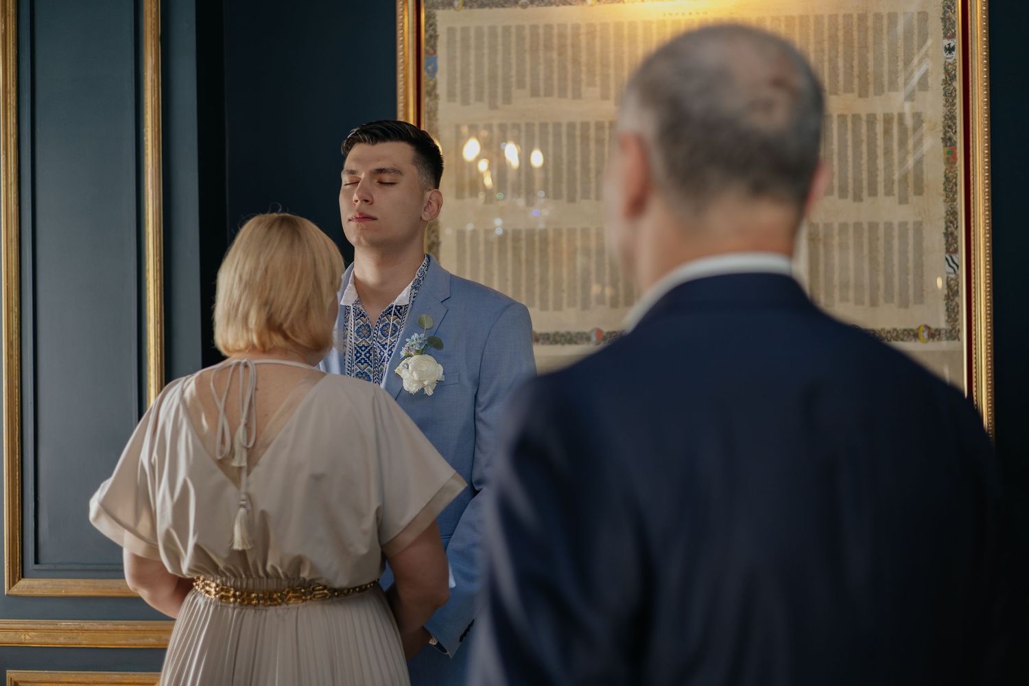 Groom in blue suit with white boutonniere stands facing bride in elegant venue during wedding ceremony.