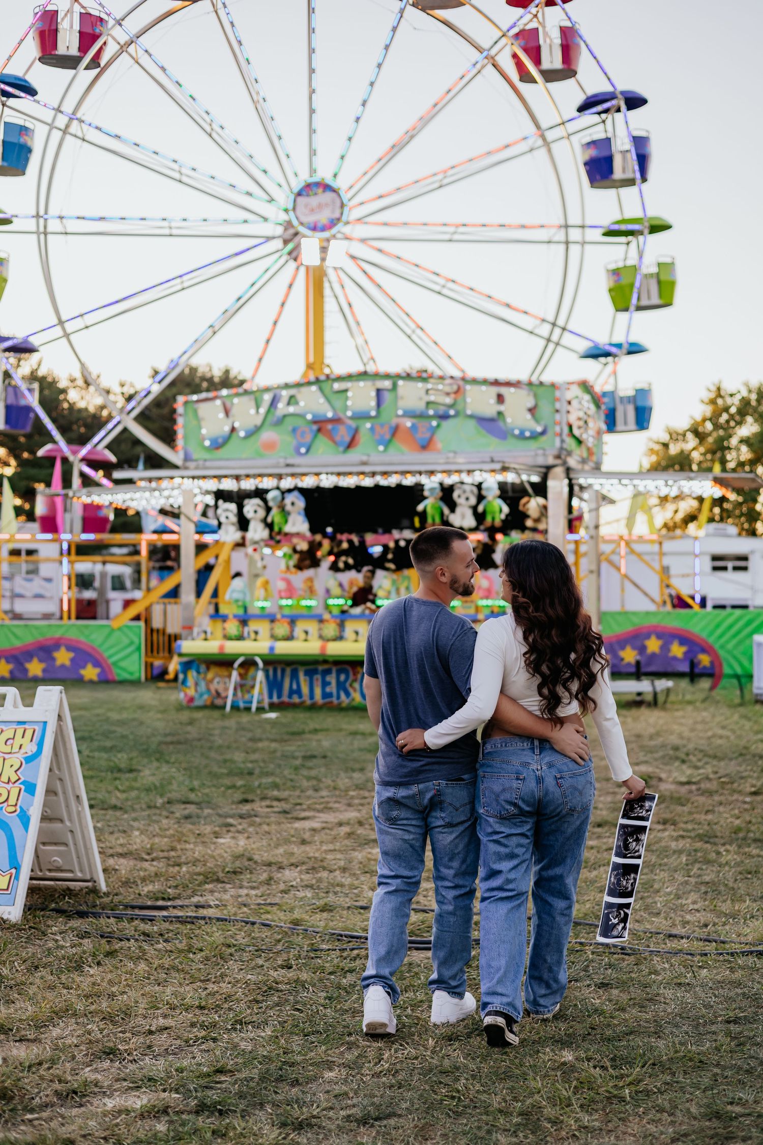 Expecting couple walking arm in arm at colorful carnival fairground with Ferris wheel in background.