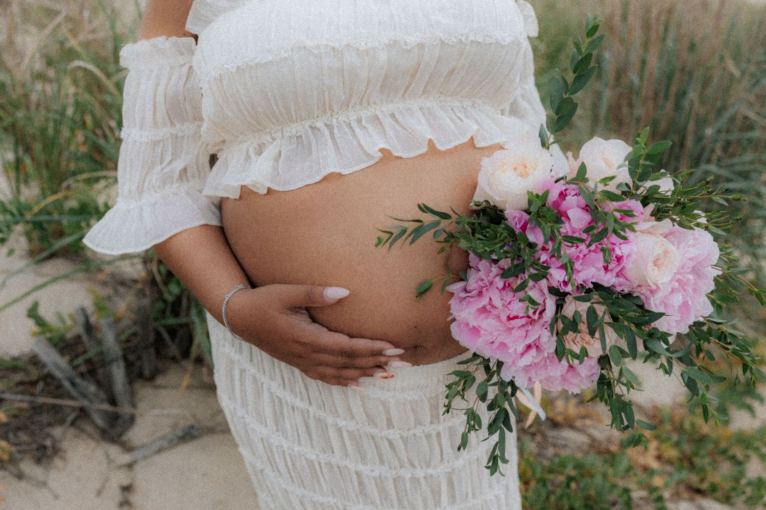 Pregnant woman in white dress holding pink peony bouquet against sandy dunes backdrop.