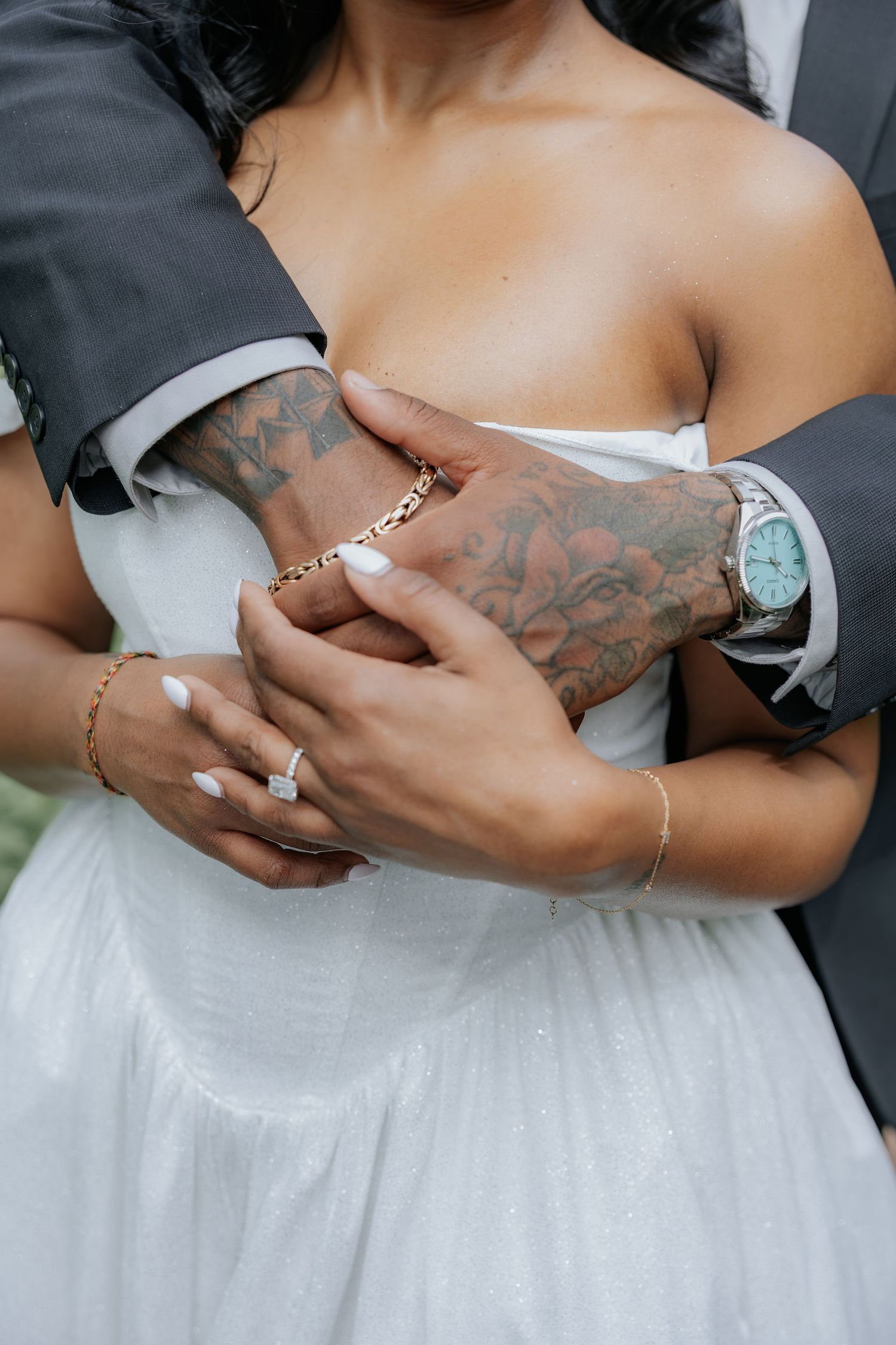 Close-up of groom's tattooed hands embracing bride's waist showing engagement ring and gold jewelry.