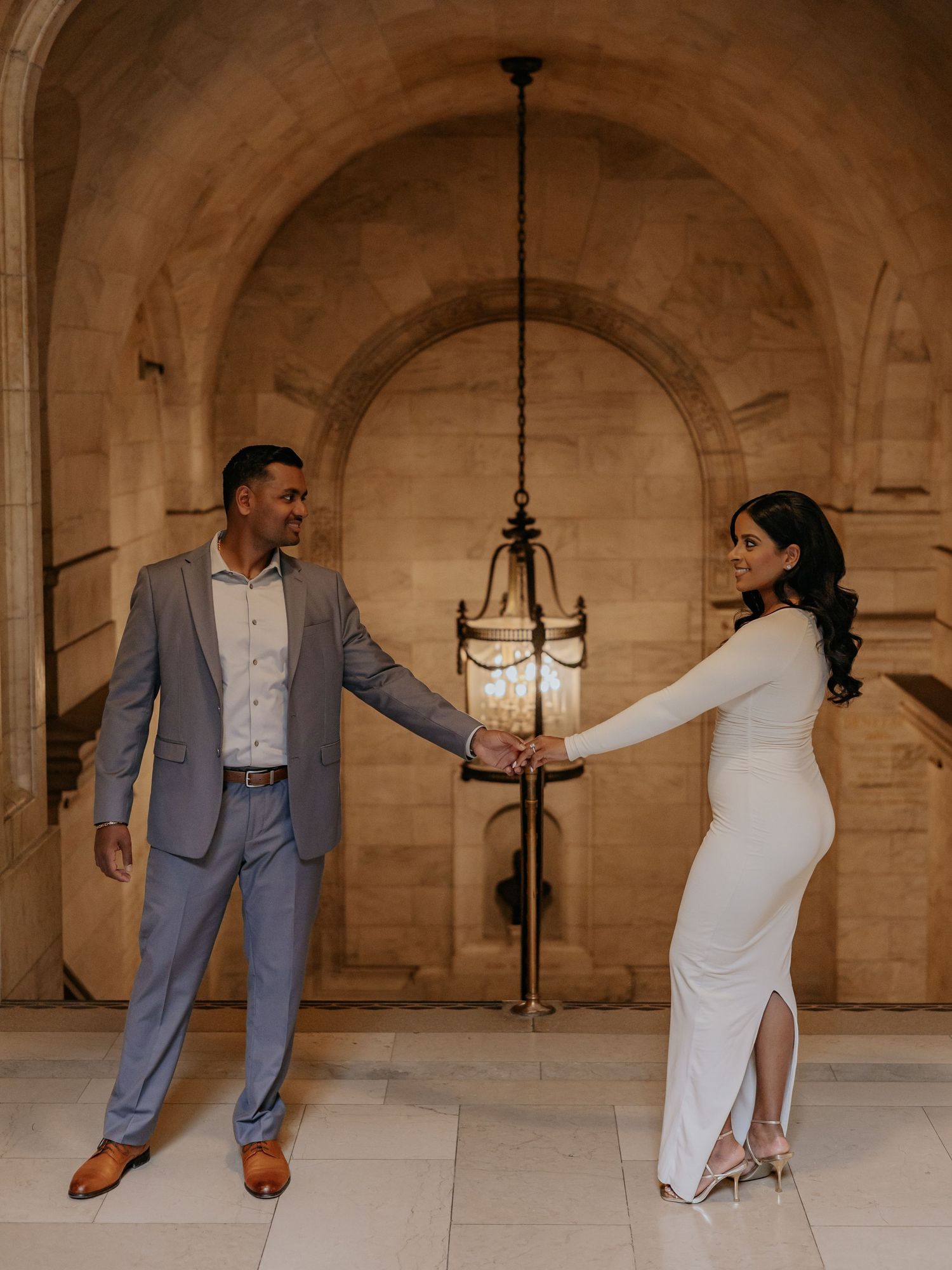 Couple holds hands facing each other under stone arch with hanging lantern.