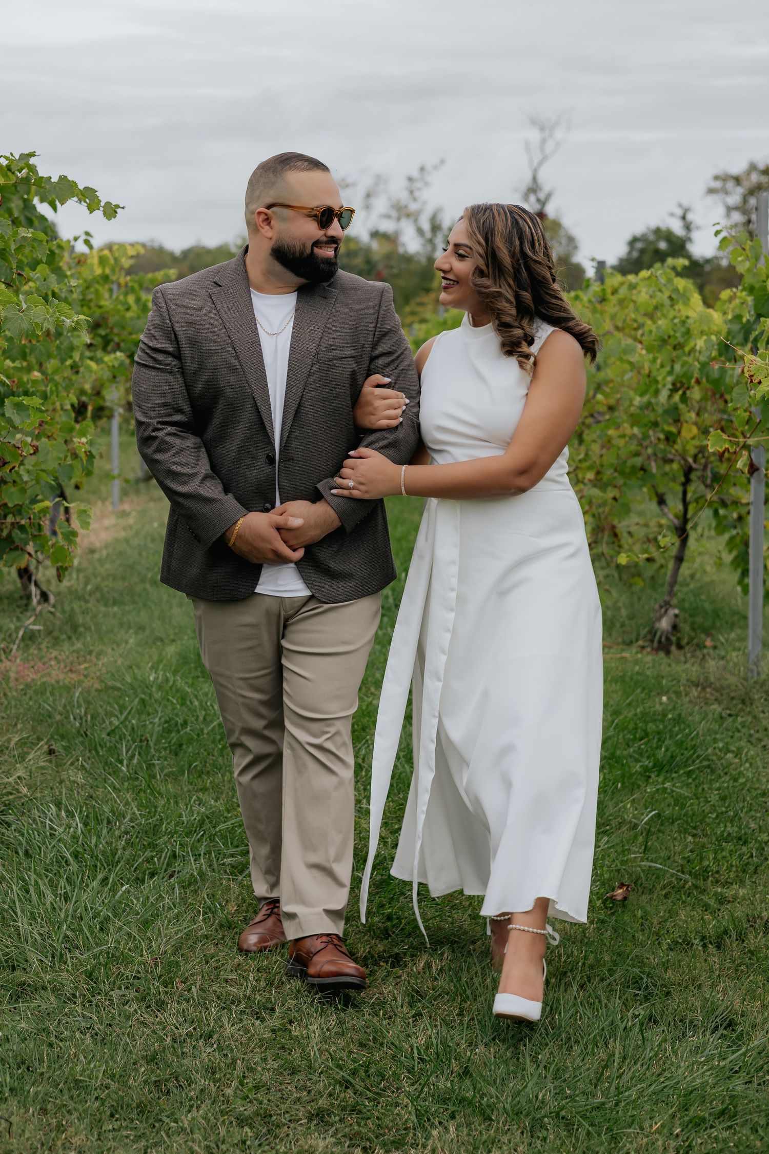 Happy couple smiling at each other in a vineyard, woman in white dress and man in brown blazer.