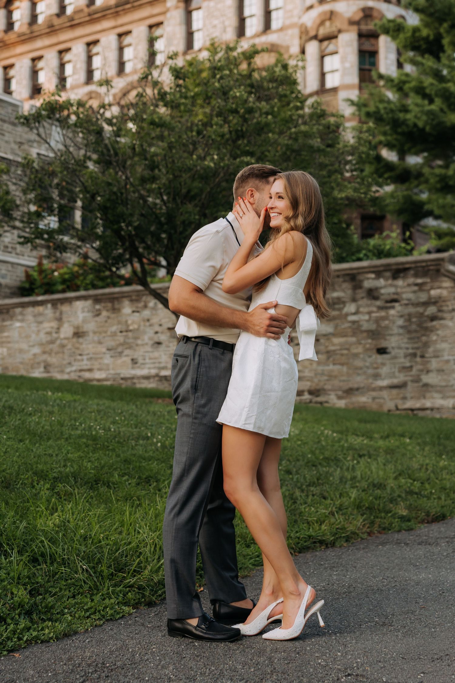 A couple embraces and kisses outdoors near a historic stone building, woman in white dress and heels.