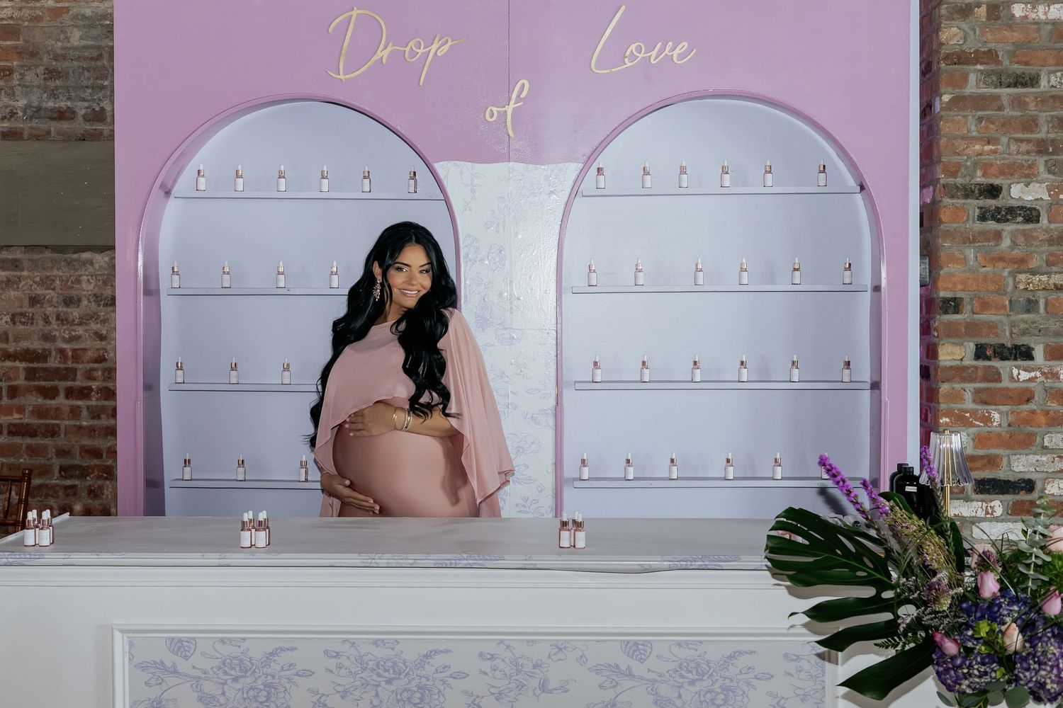 Pregnant woman in pink maternity gown standing behind marble counter at Drop of Love fragrance display.