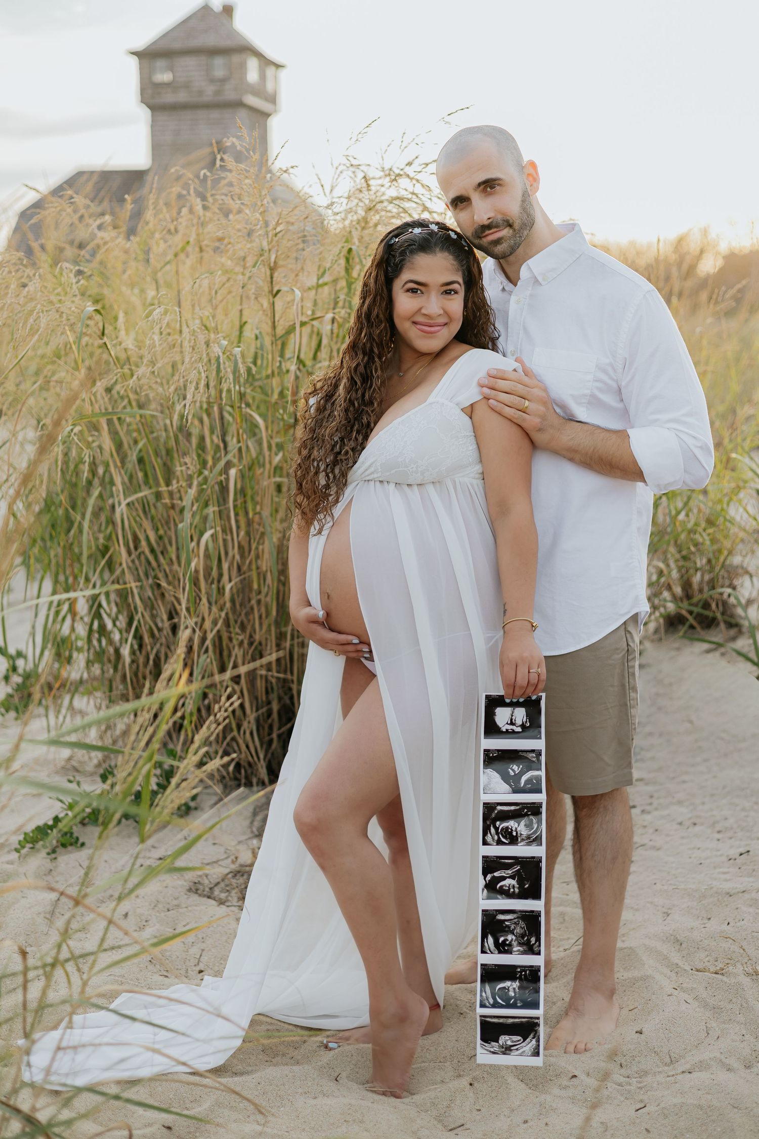 Pregnant couple poses on sandy beach dunes, woman in white maternity gown holding ultrasound photos.