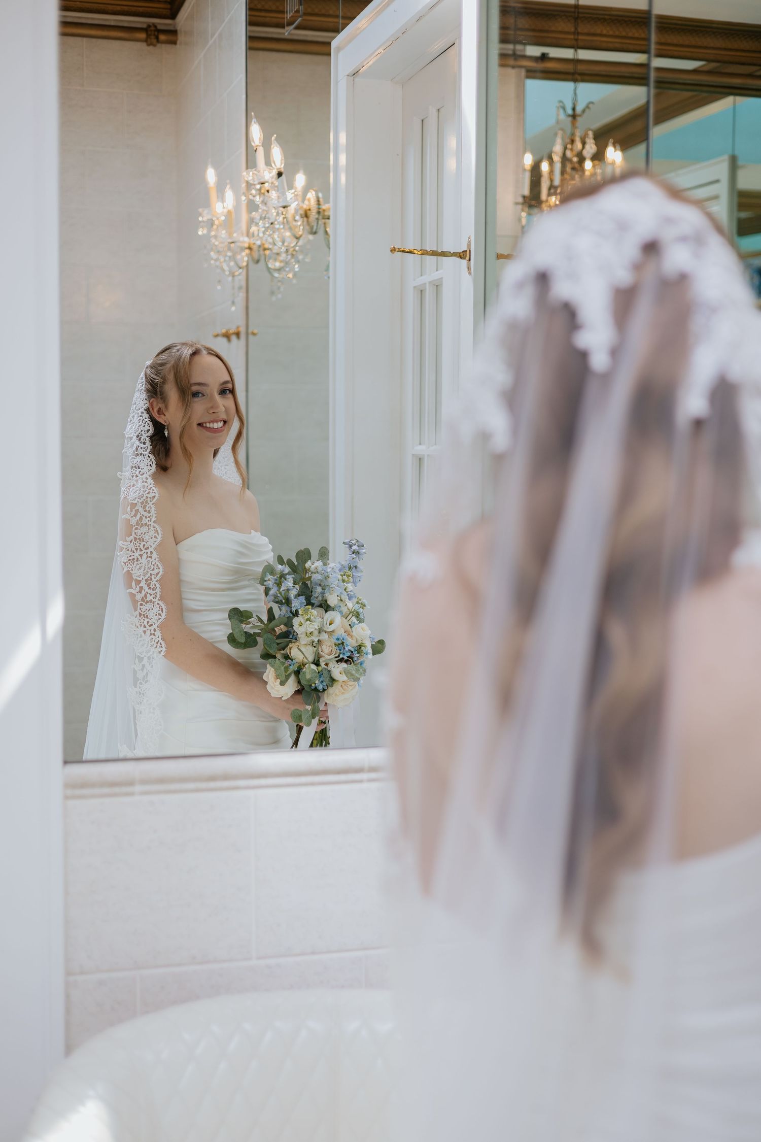 Bride in strapless gown and lace veil smiles at her reflection in ornate mirror holding floral bouquet.