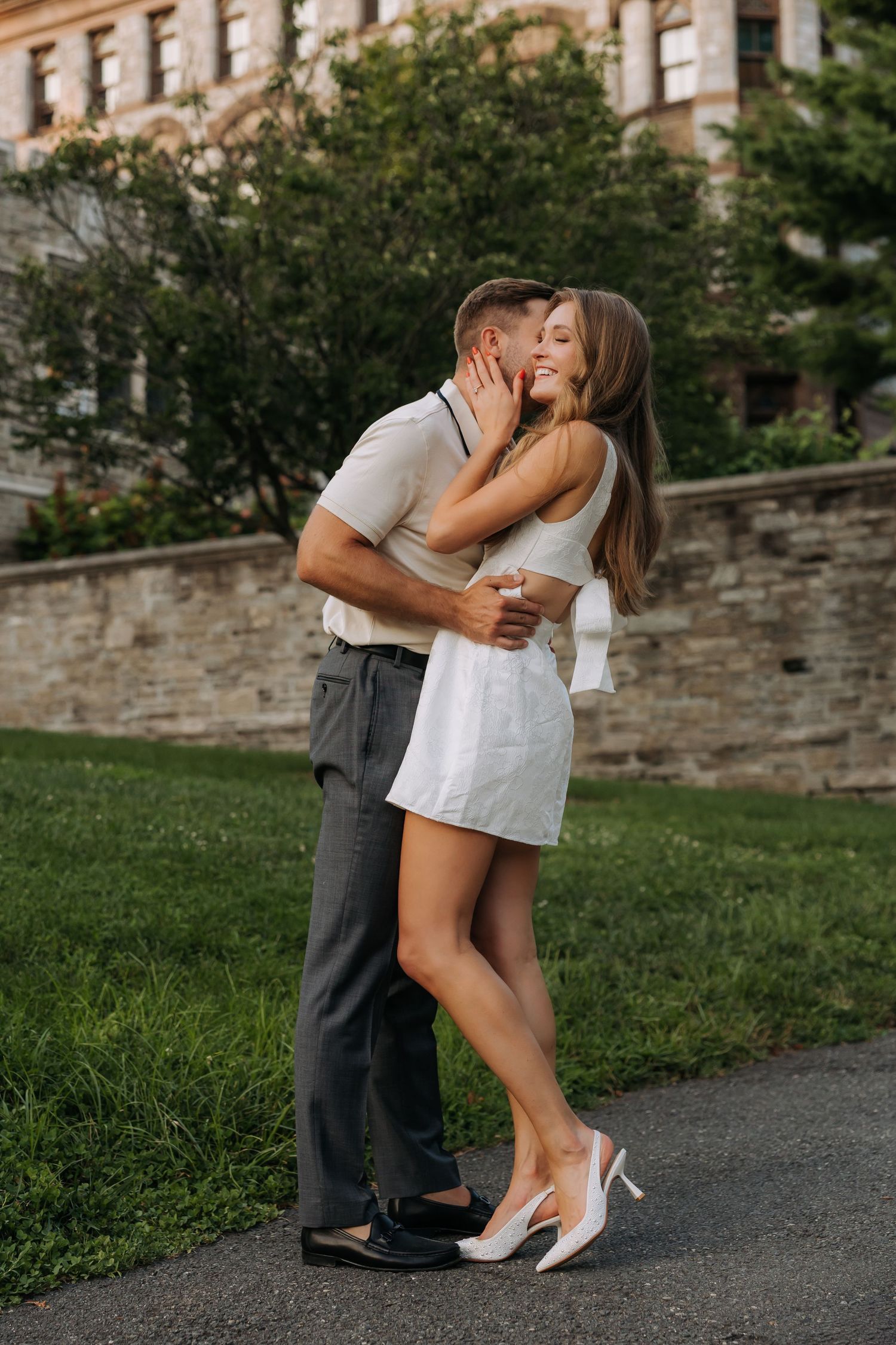 Romantic couple sharing a kiss outdoors, woman in white mini dress and heels embracing man.