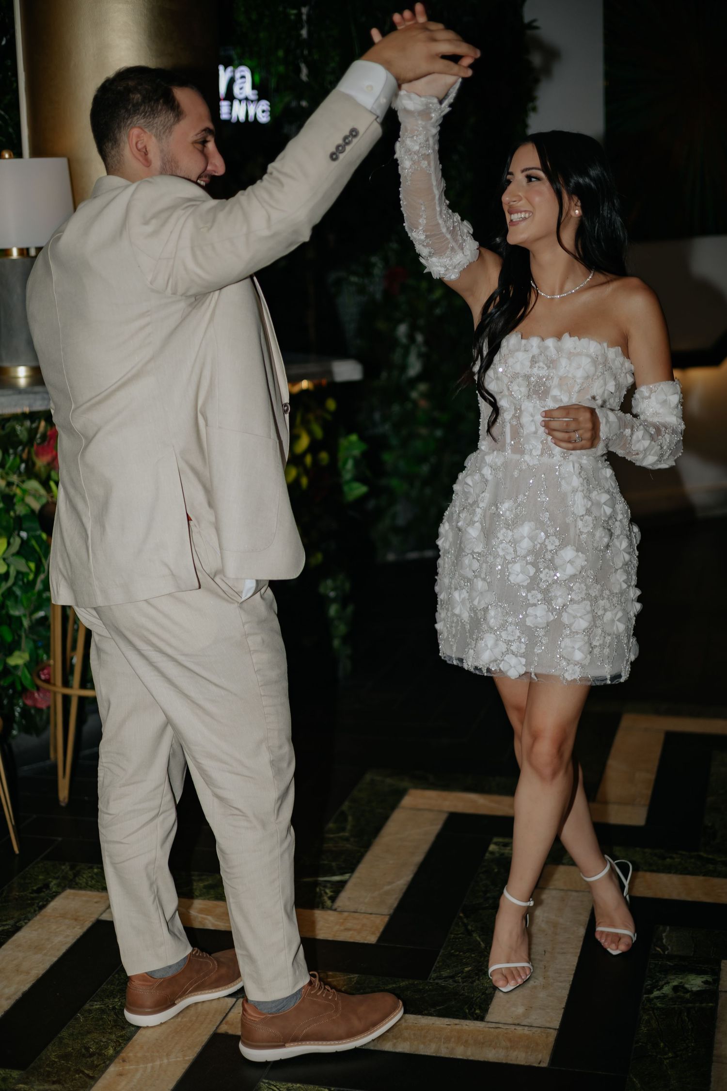 Newlyweds dance joyfully, bride in a short white floral dress, groom in a cream suit at their reception.