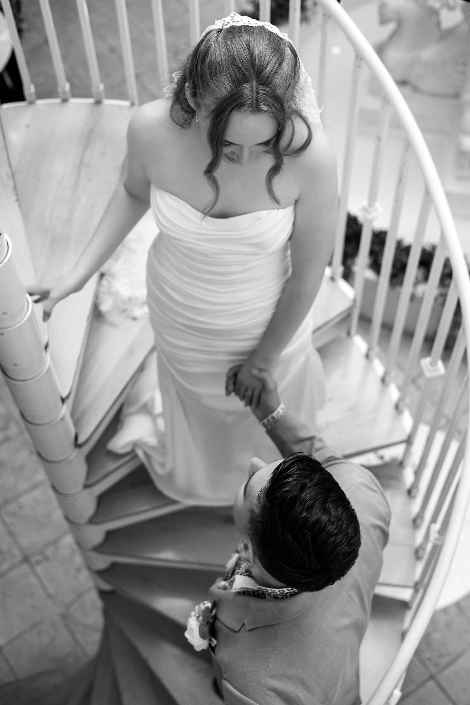 Black and white overhead view of bride in white gown and groom holding hands on a spiral staircase.