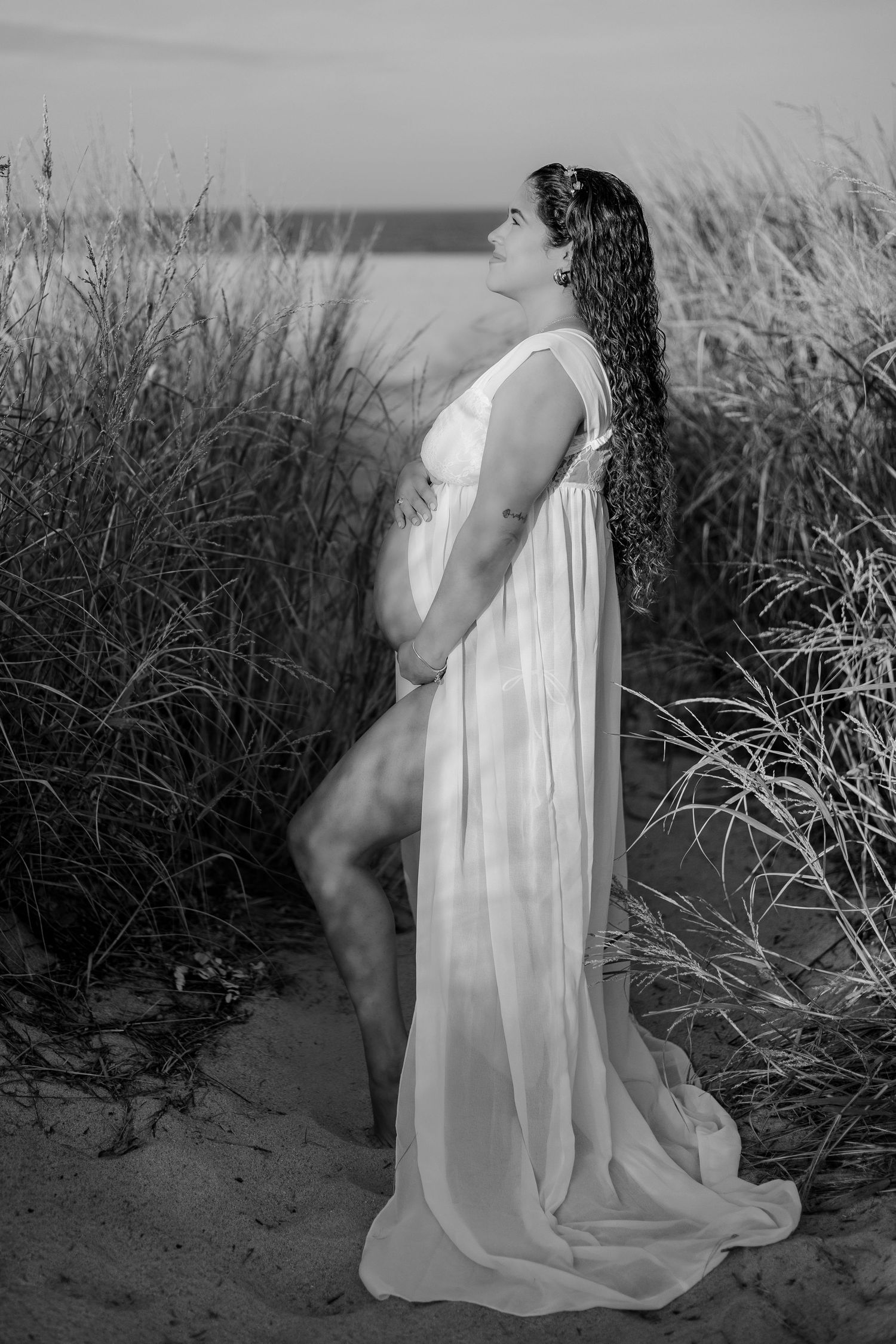 Black and white photo of pregnant woman in flowing white sheer gown standing among tall beach grass at dusk.