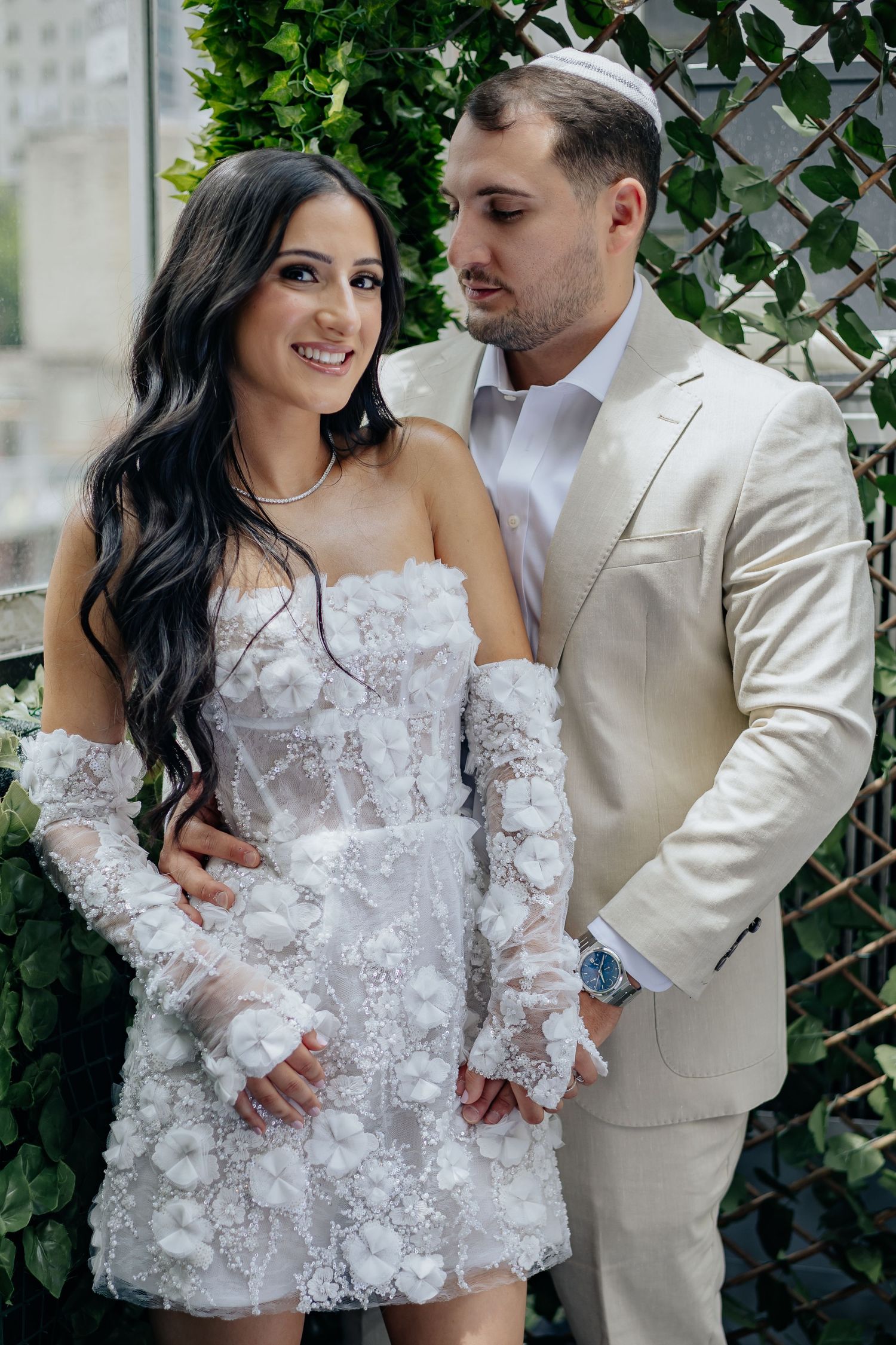 Elegant bride in floral applique mini dress stands with groom in cream suit at wedding.