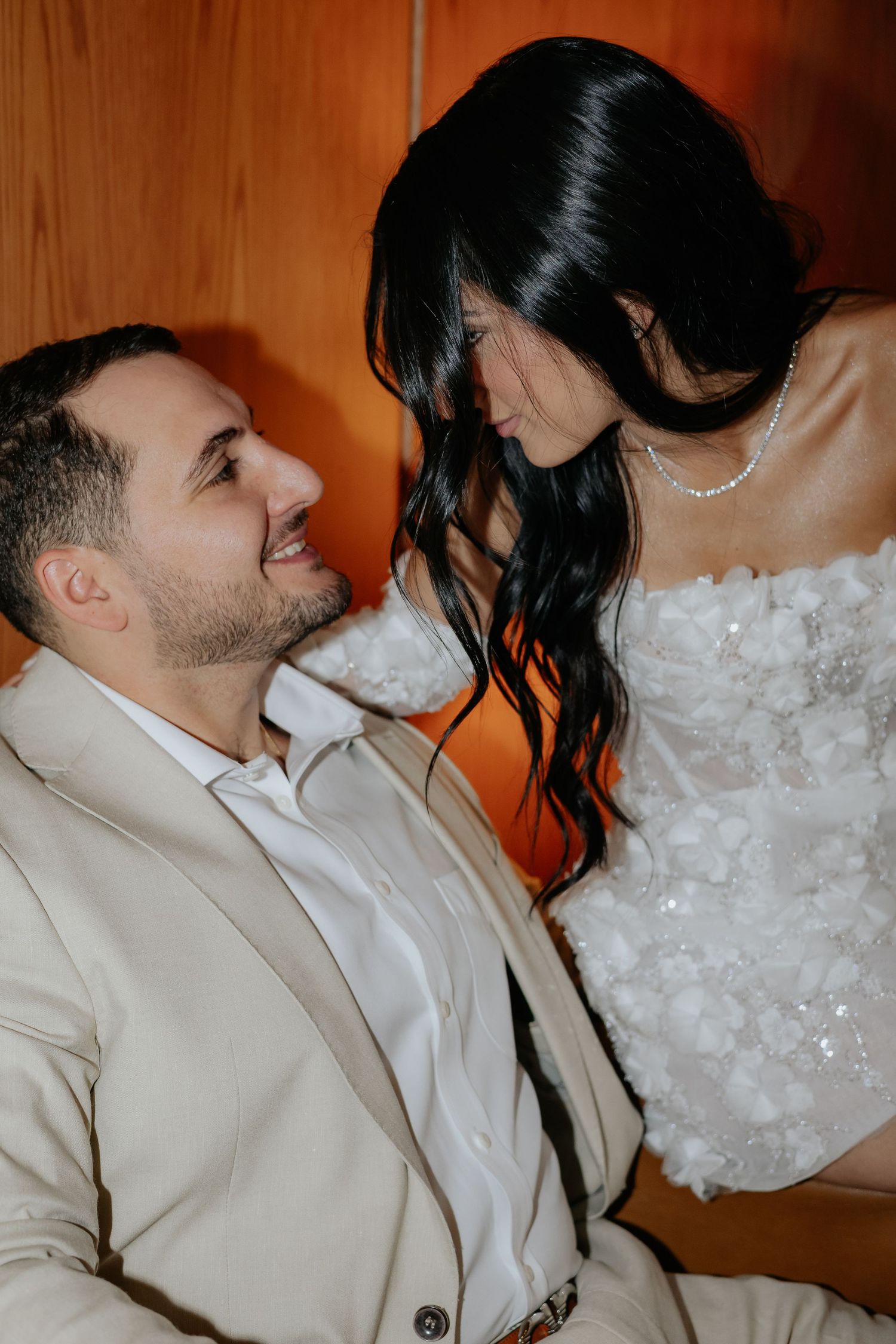 Groom smiling up at bride as they sit together against a warm wooden background.