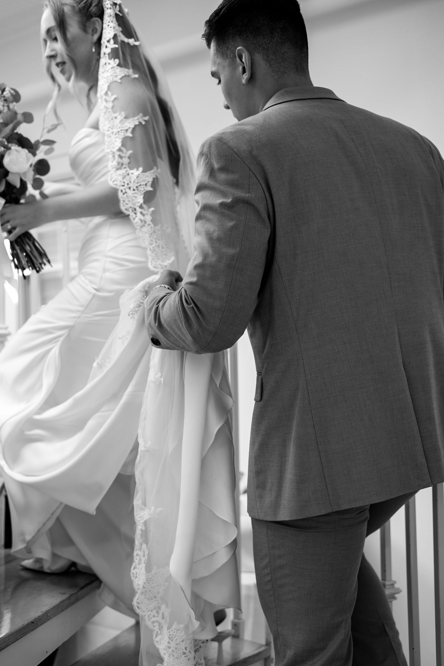 Groom carrying bride's train up stairs as she holds bouquet, black and white wedding photo.