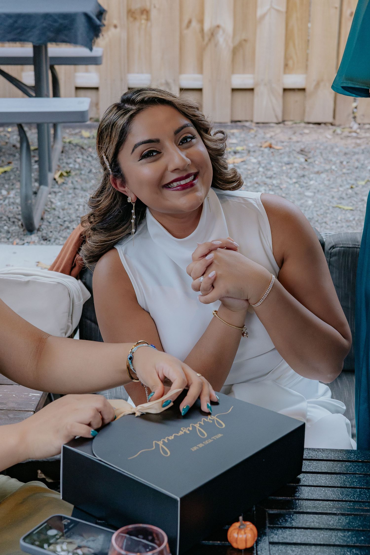 Smiling woman in white dress opening a gift box at an outdoor celebration.