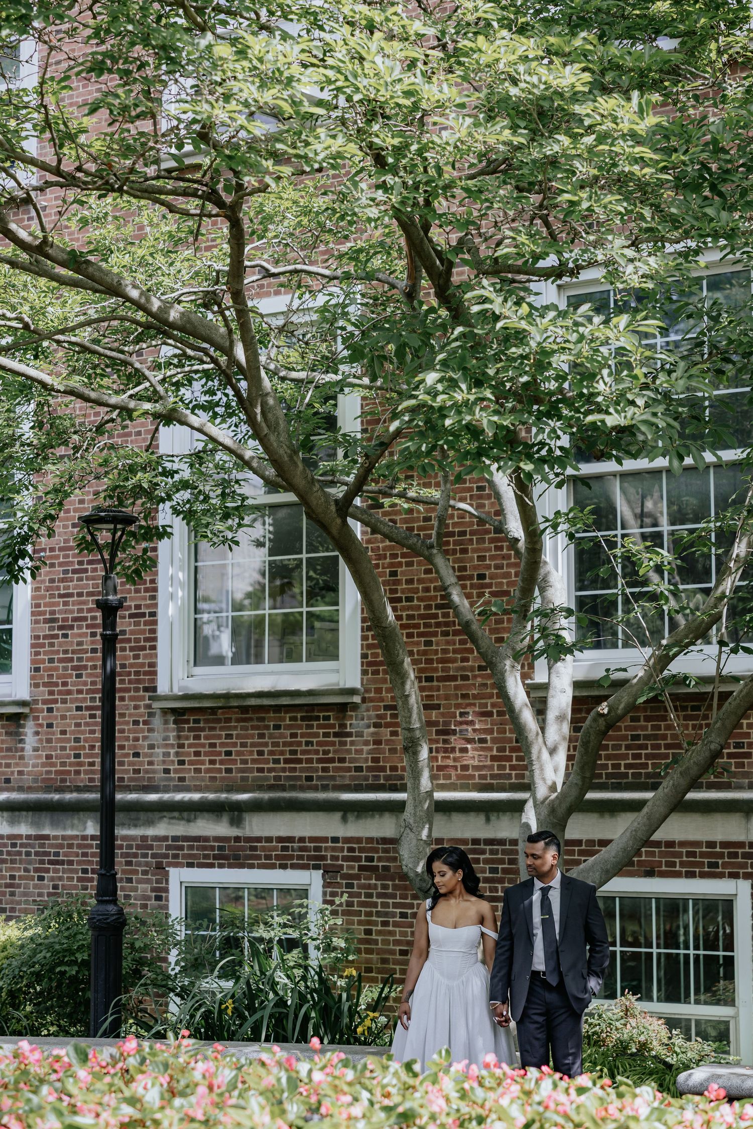 Bride and groom walk hand in hand through a garden with a brick building backdrop.