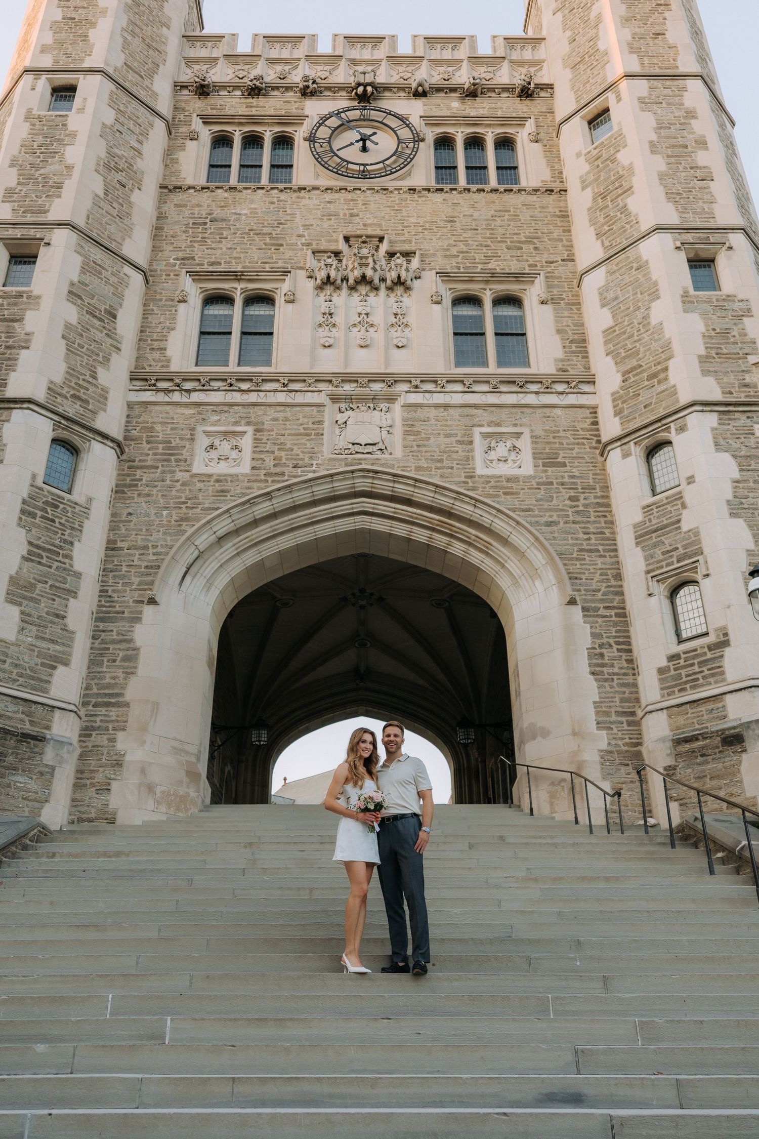 Couple posing on stone steps beneath Gothic arch of historic university building with clock tower.