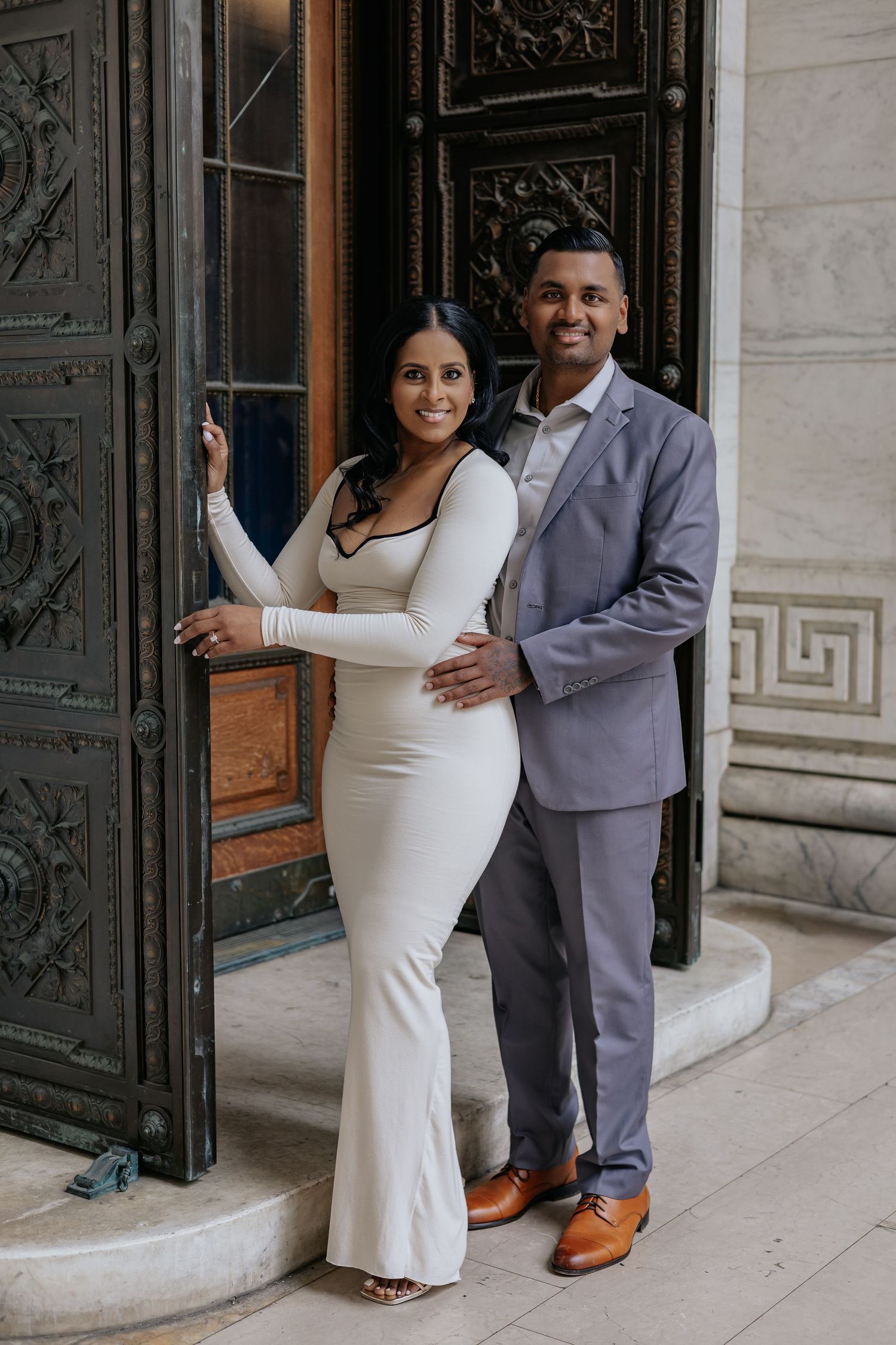 Stylish engaged couple posing by ornate bronze doors of a classic stone building.