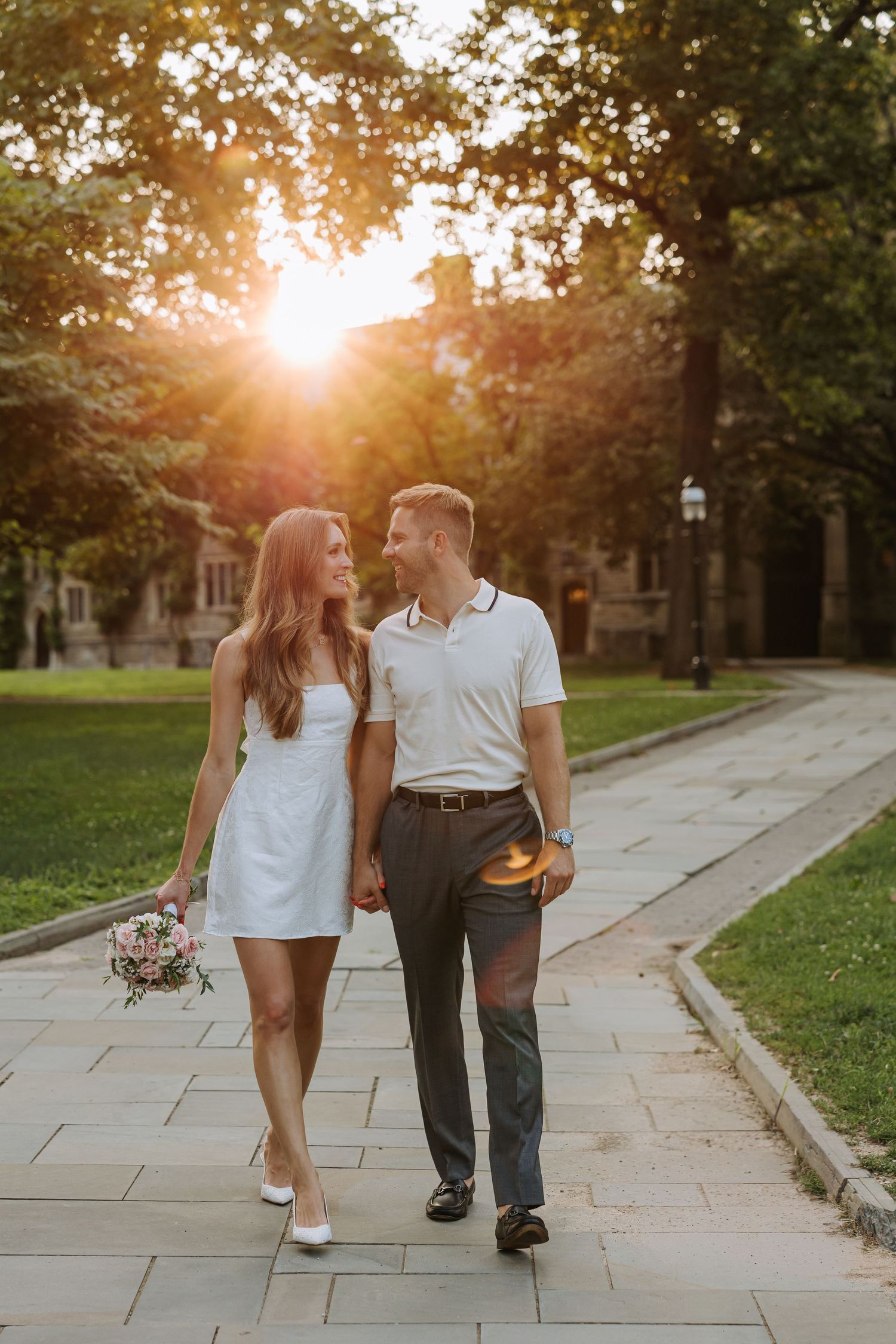 Couple holding hands walking on a sunlit pathway, woman in white dress with bouquet, man in white polo and gray pants.