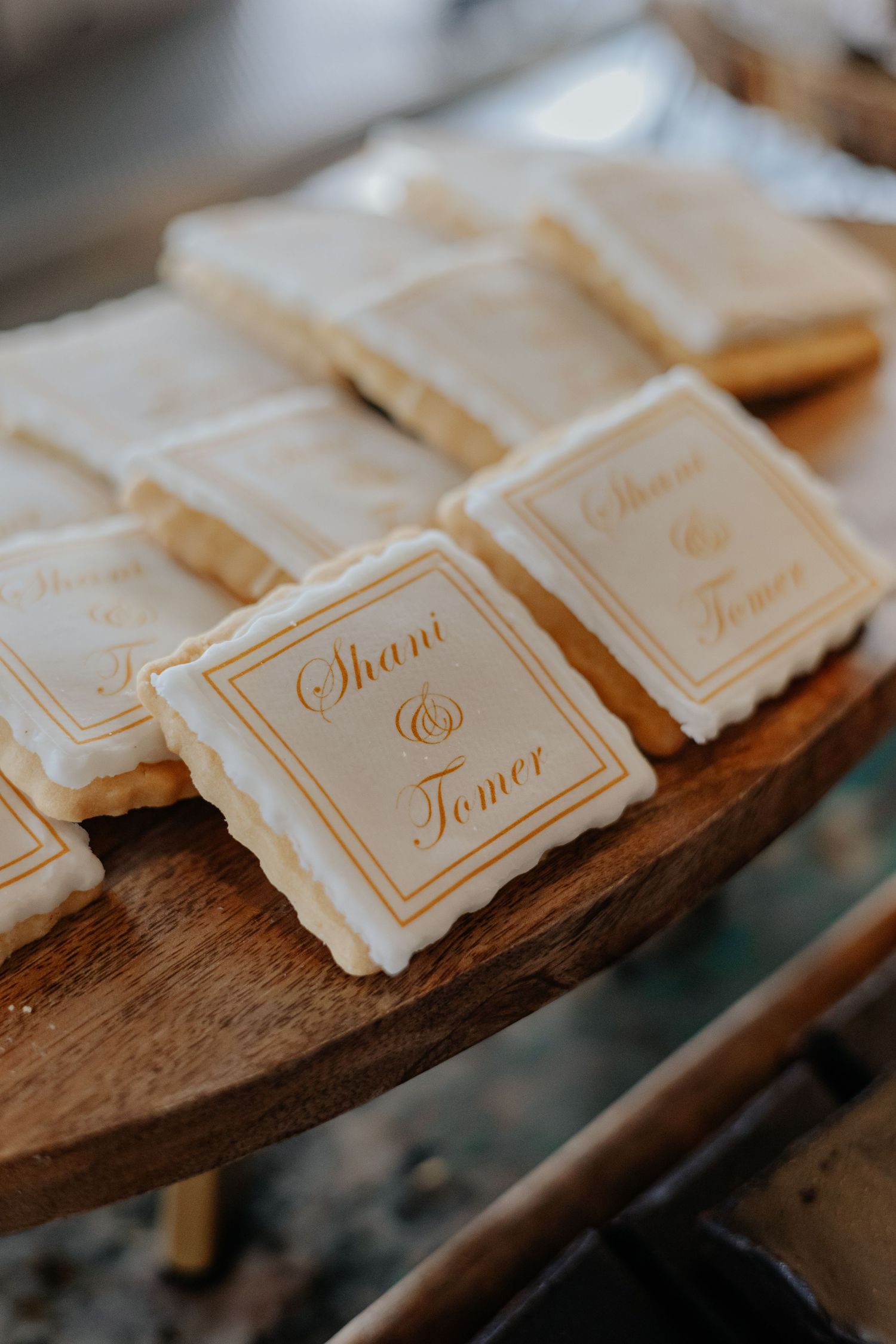 Custom white and gold wedding cookies reading Shani and Tomer displayed on a wooden cake stand.