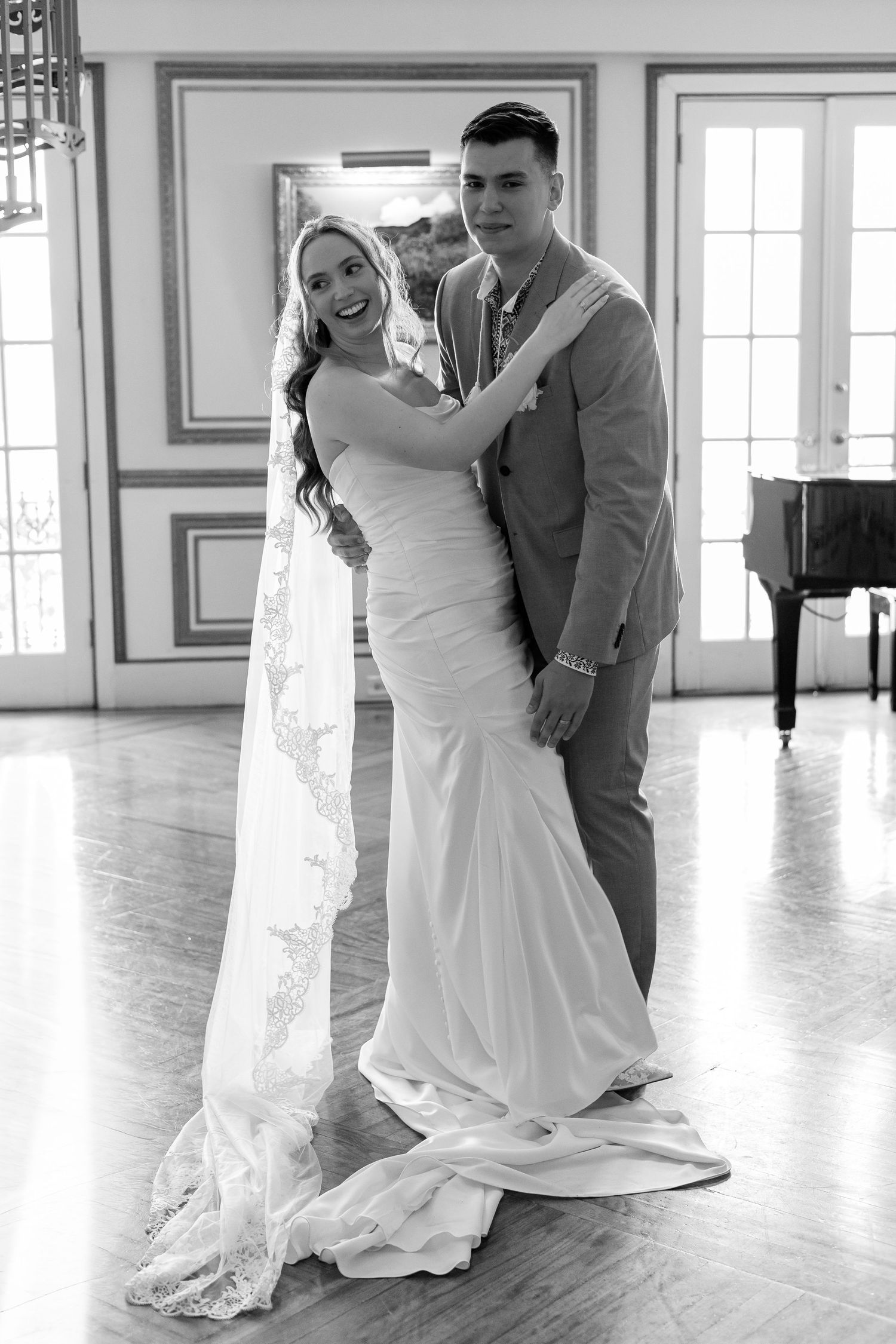 Bride and groom posing in elegant ballroom, bride in fitted gown with long lace veil, black and white.