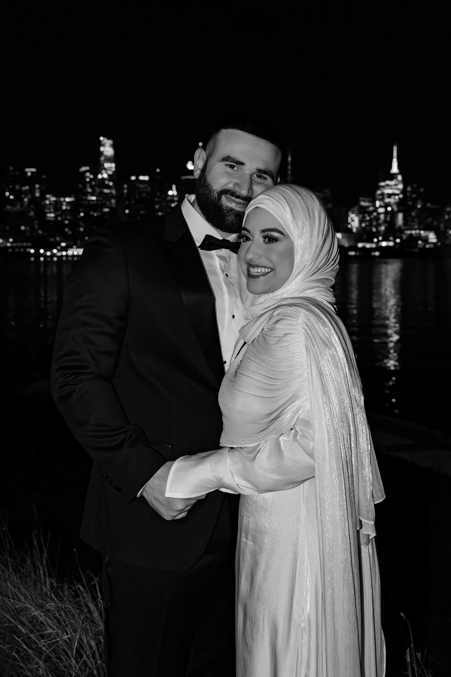 Black and white portrait of smiling Muslim bride and groom embracing with city skyline at night.