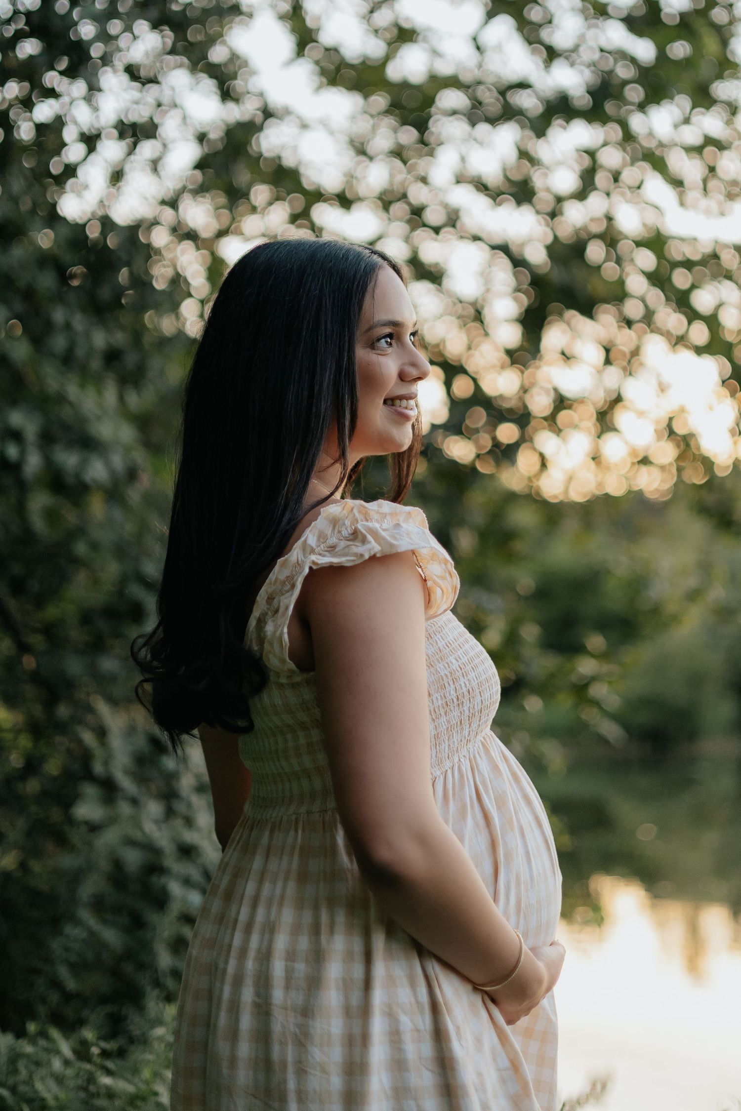 Radiant pregnant woman in yellow gingham dress smiling over shoulder in golden hour light near a serene pond.