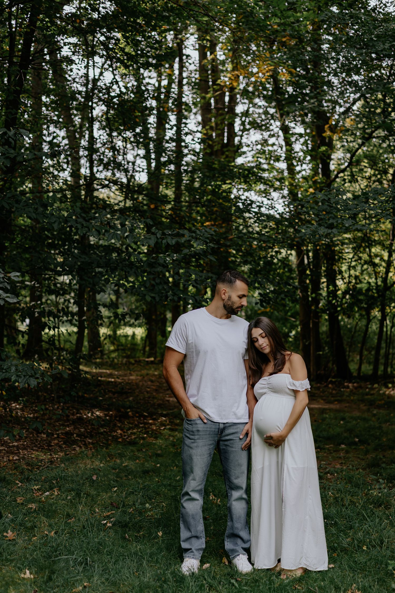 Expecting couple standing together in a lush green forest, woman in white maternity dress cradling her baby bump.