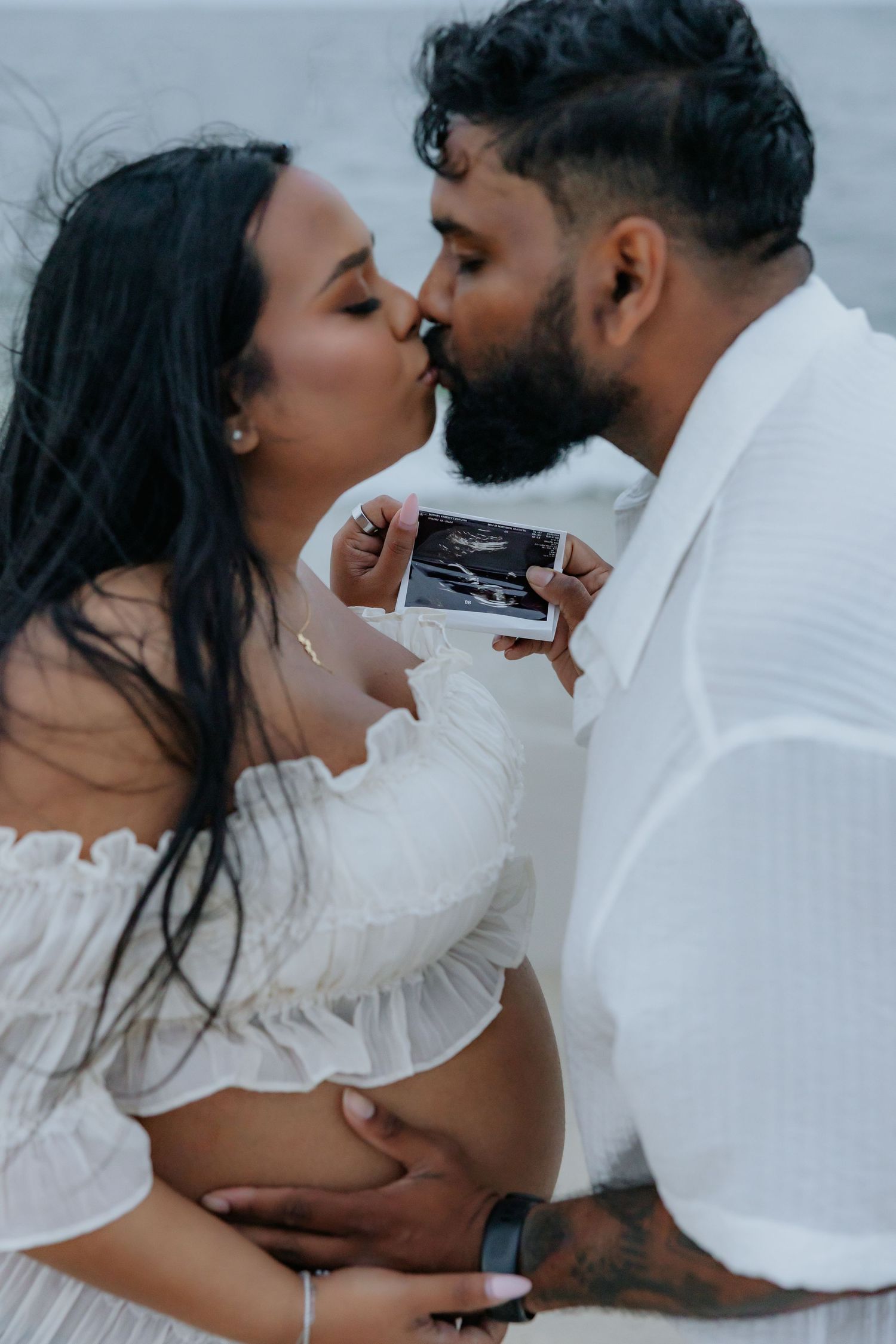 Expecting couple kissing while holding ultrasound photo during romantic outdoor maternity photoshoot.