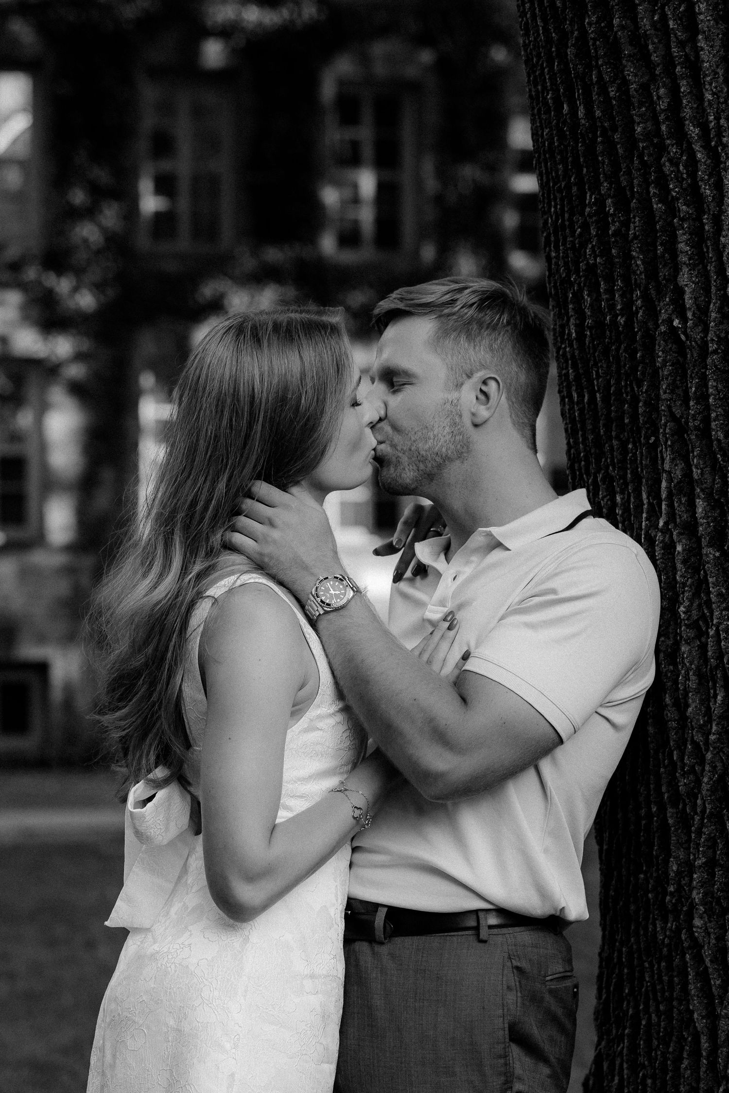 Black and white photo of a couple sharing a romantic kiss near a tree, with a historic building in the background.