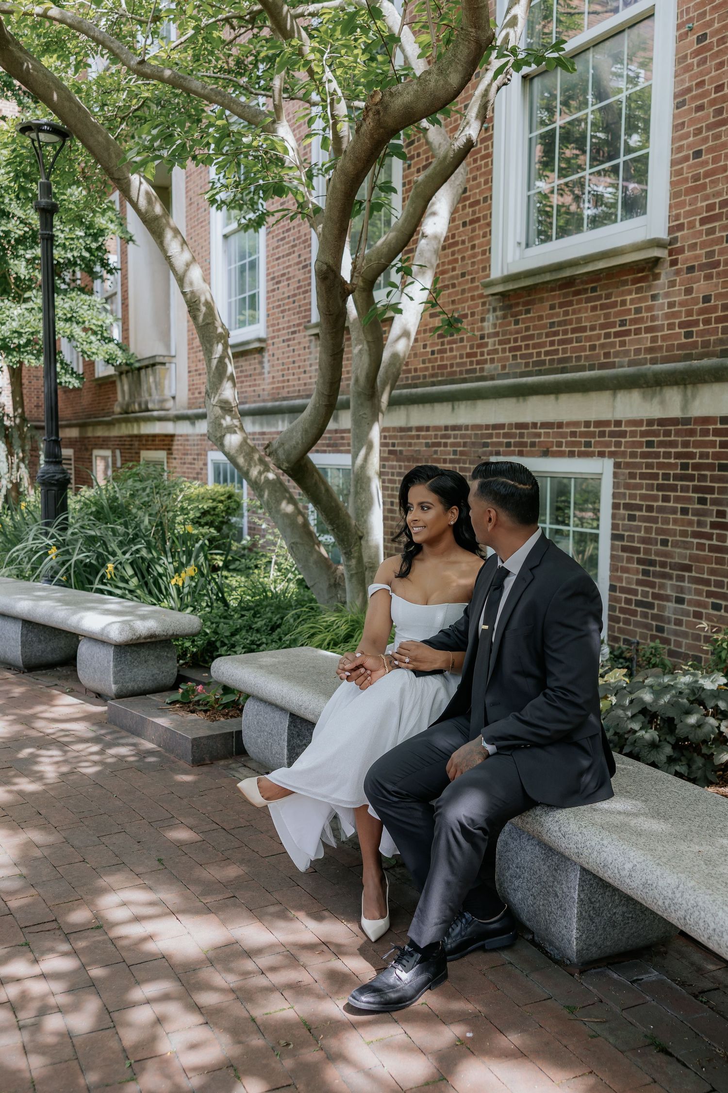 Bride in white dress and groom in dark suit sitting together on stone bench outdoors.
