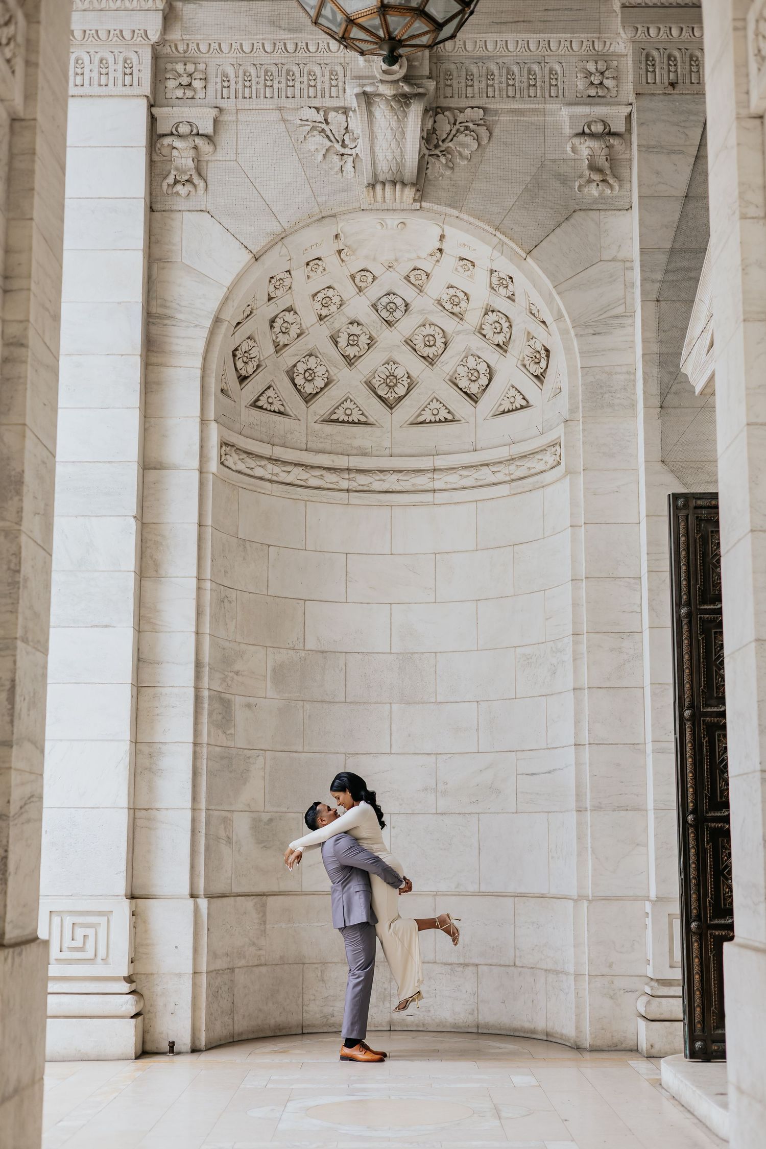 Groom lifts bride in white gown beneath ornate carved marble archway.