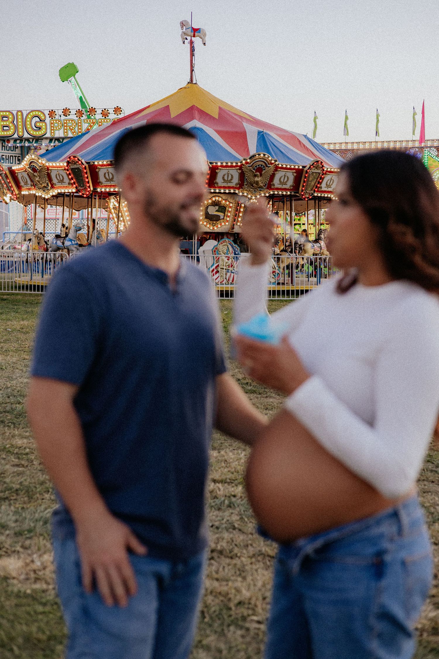 Pregnant couple standing near illuminated carousel at a county fair.