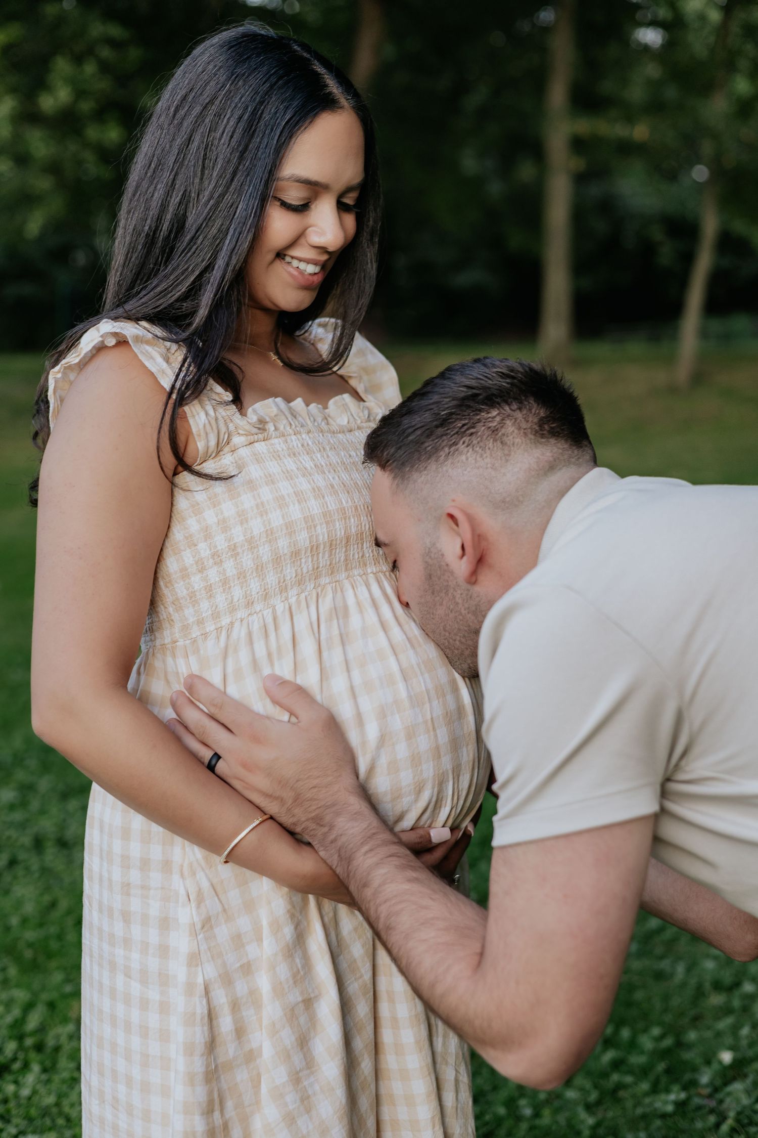 Man lovingly kissing pregnant woman's baby bump while she smiles down at him in a lush green park.