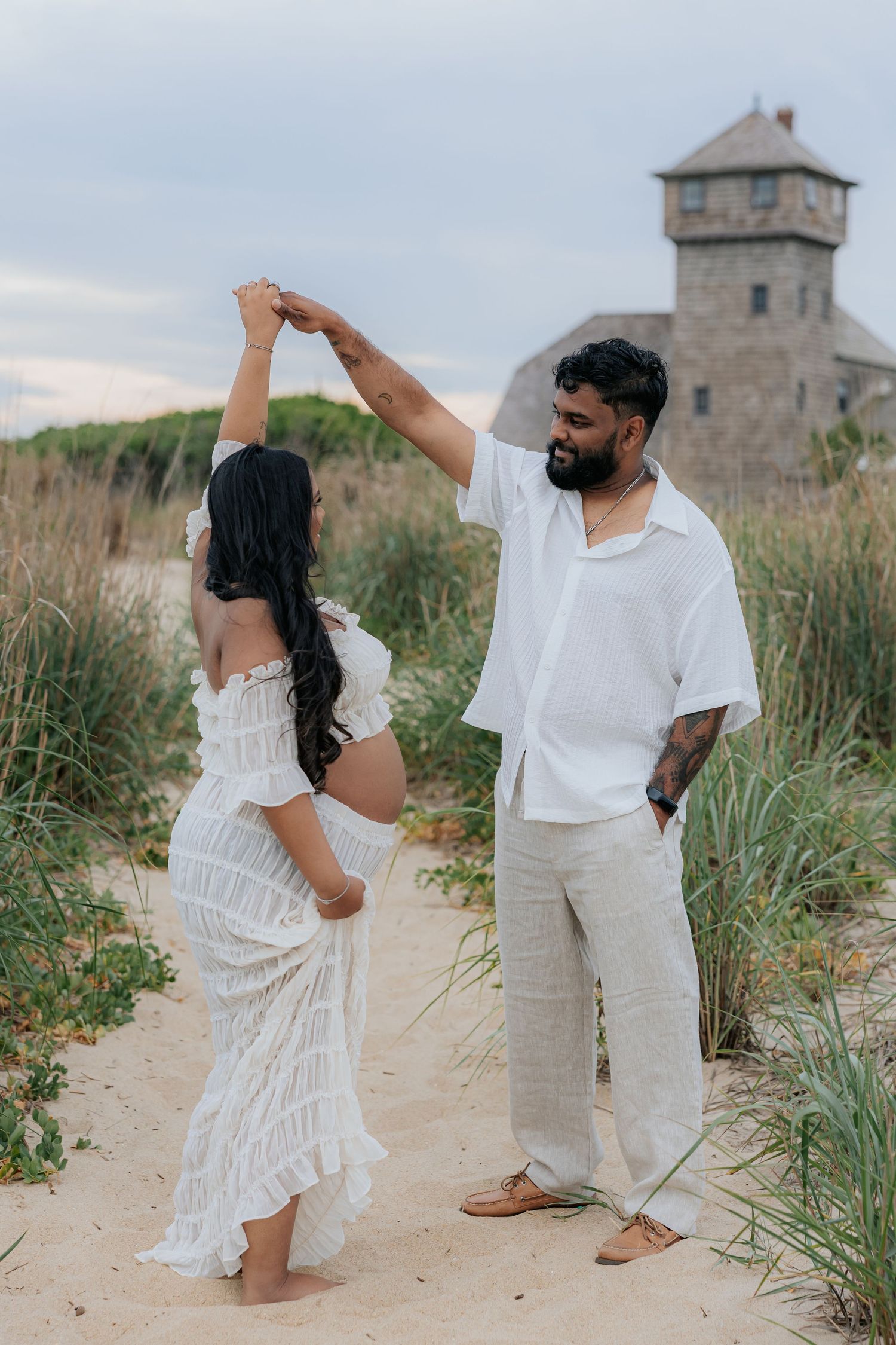 Couple dancing on sandy beach dunes near historic stone tower at dusk.