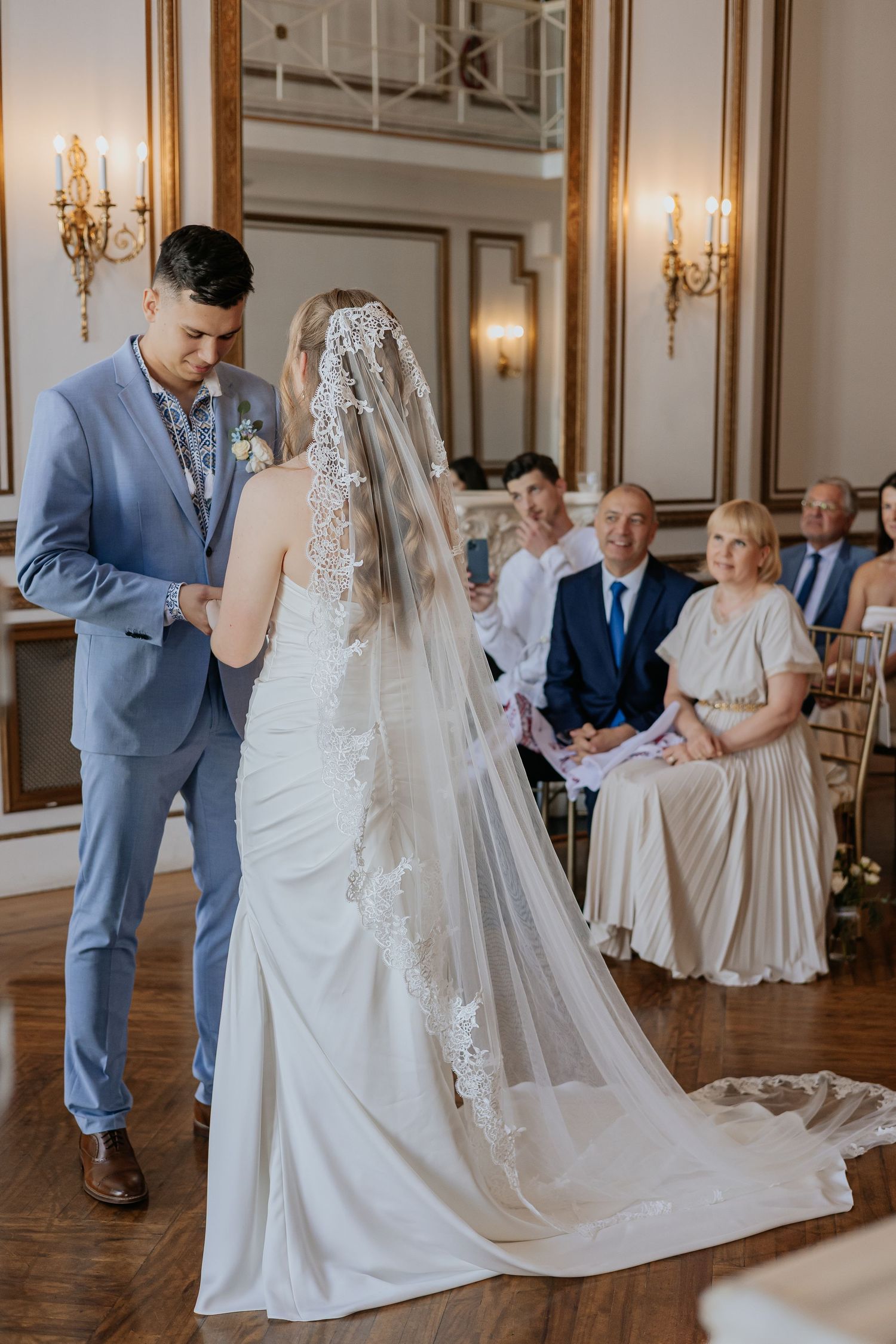 Groom in blue suit lifts bride's lace veil during wedding ceremony as guests watch from seats.