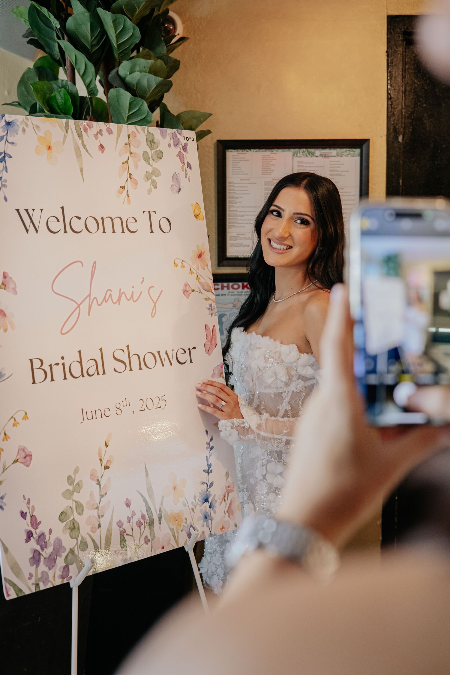 Smiling bride-to-be posing beside a floral Welcome to Shani's Bridal Shower sign.