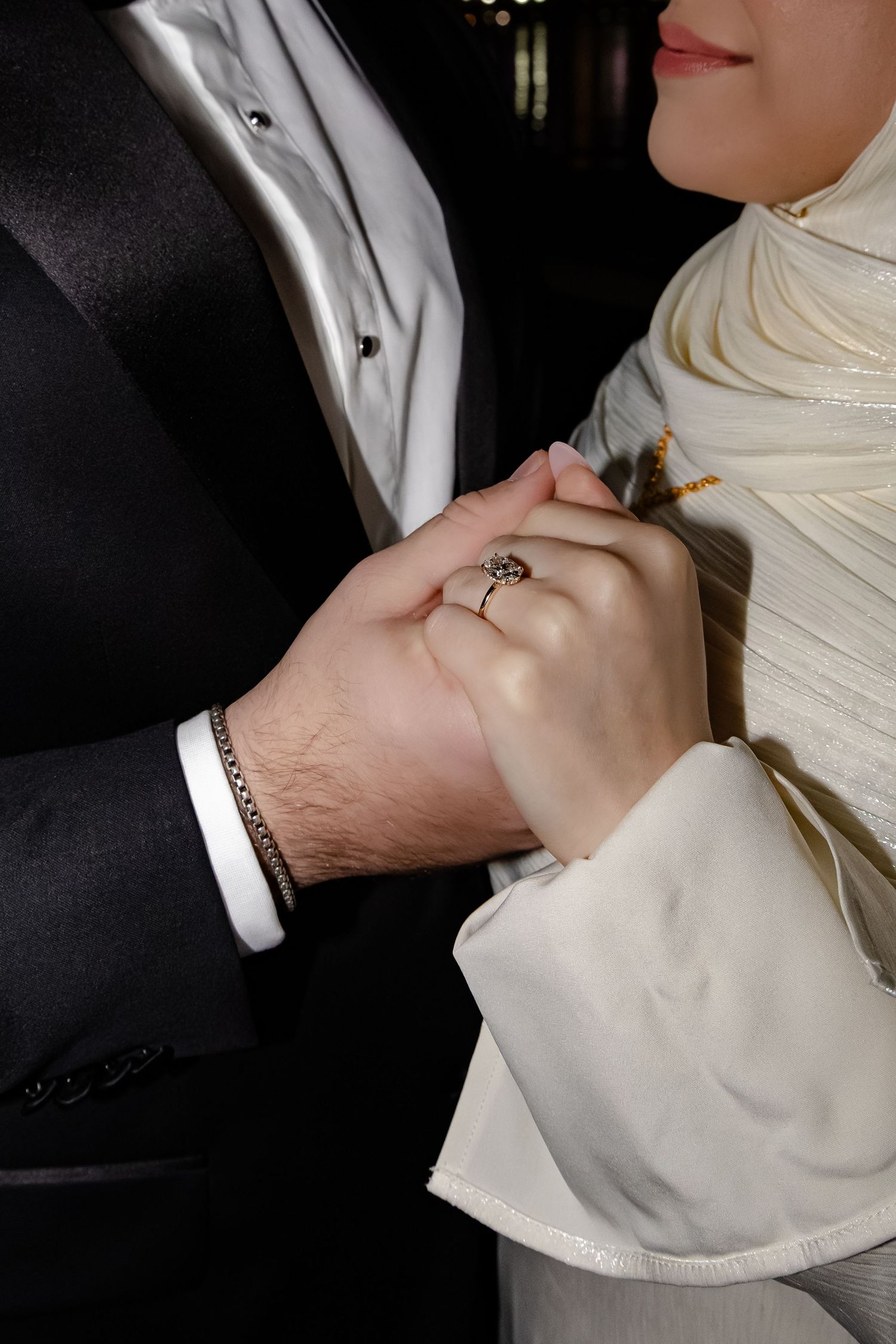 Close-up of bride and groom holding hands, showing wedding ring and bracelet in formal attire.