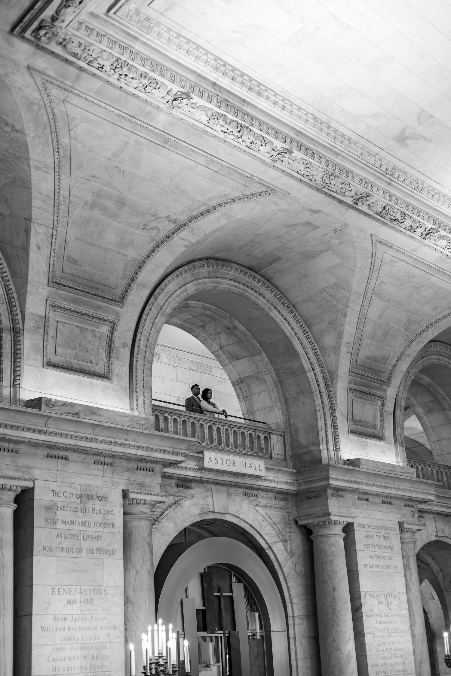 Black and white photo of couple on balcony framed by soaring marble arched hall.
