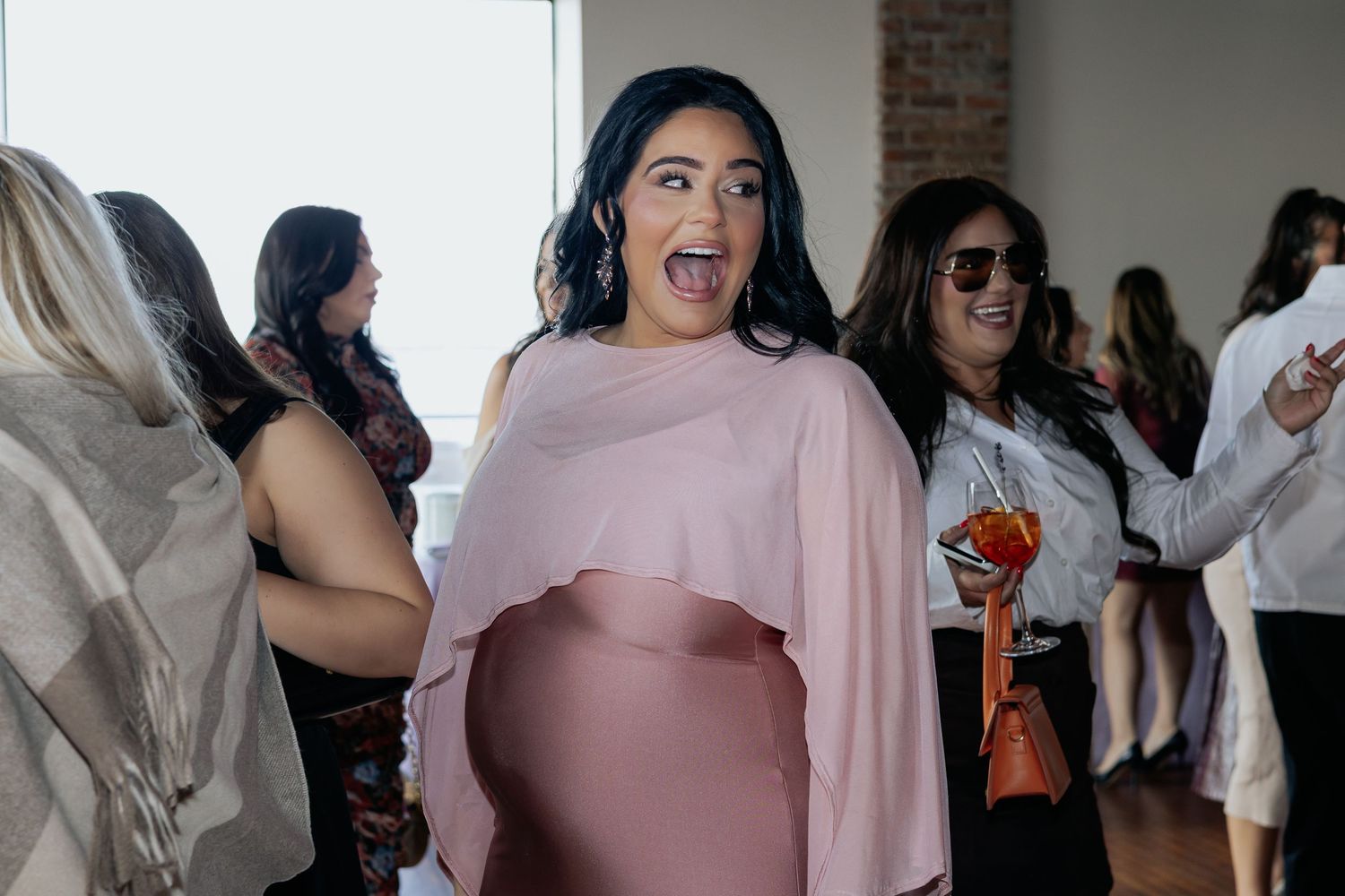 Excited pregnant woman in pink dress with surprised expression at a baby shower celebration.