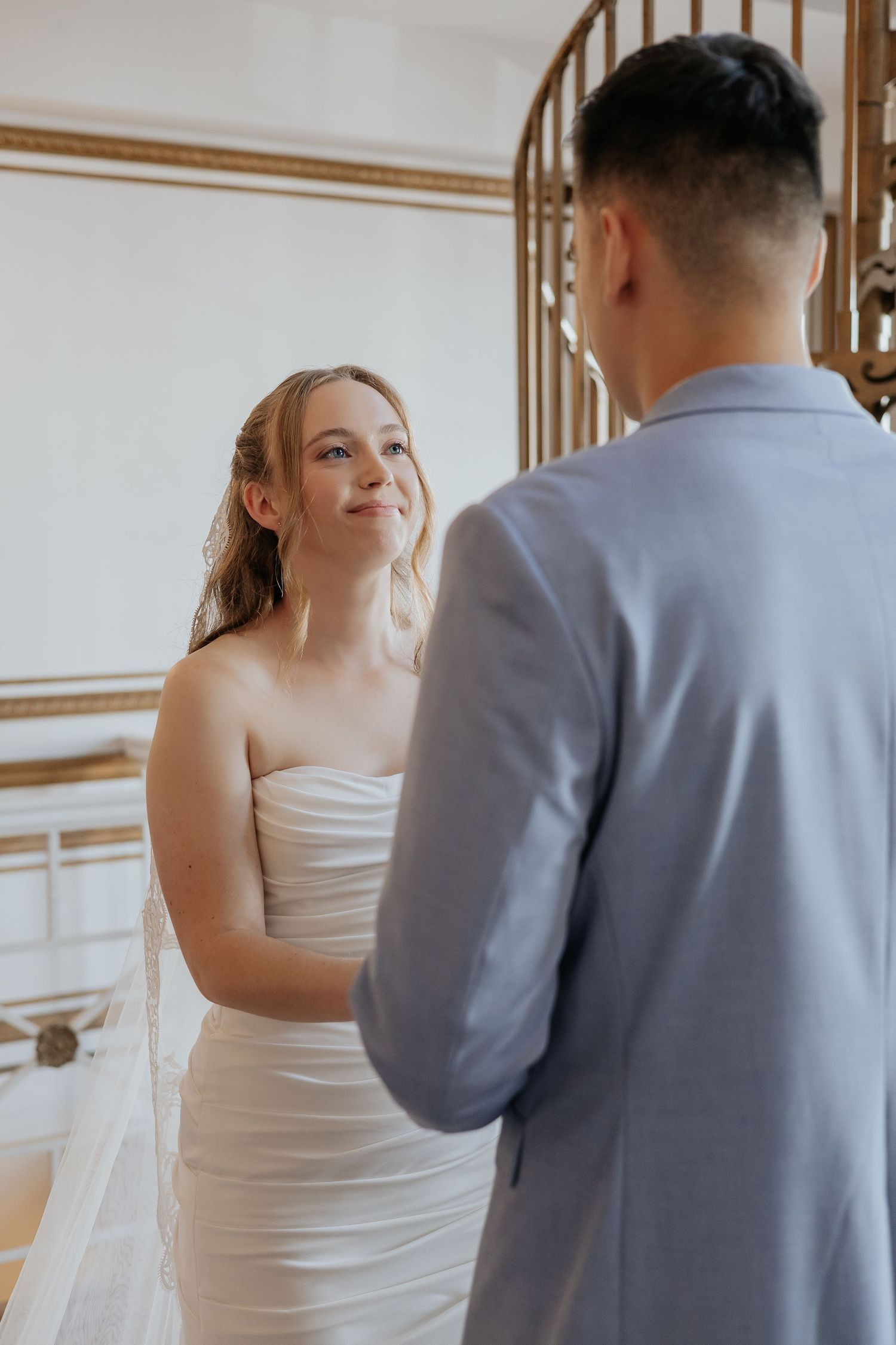 Smiling bride in strapless white gown gazes at groom during intimate indoor wedding ceremony.