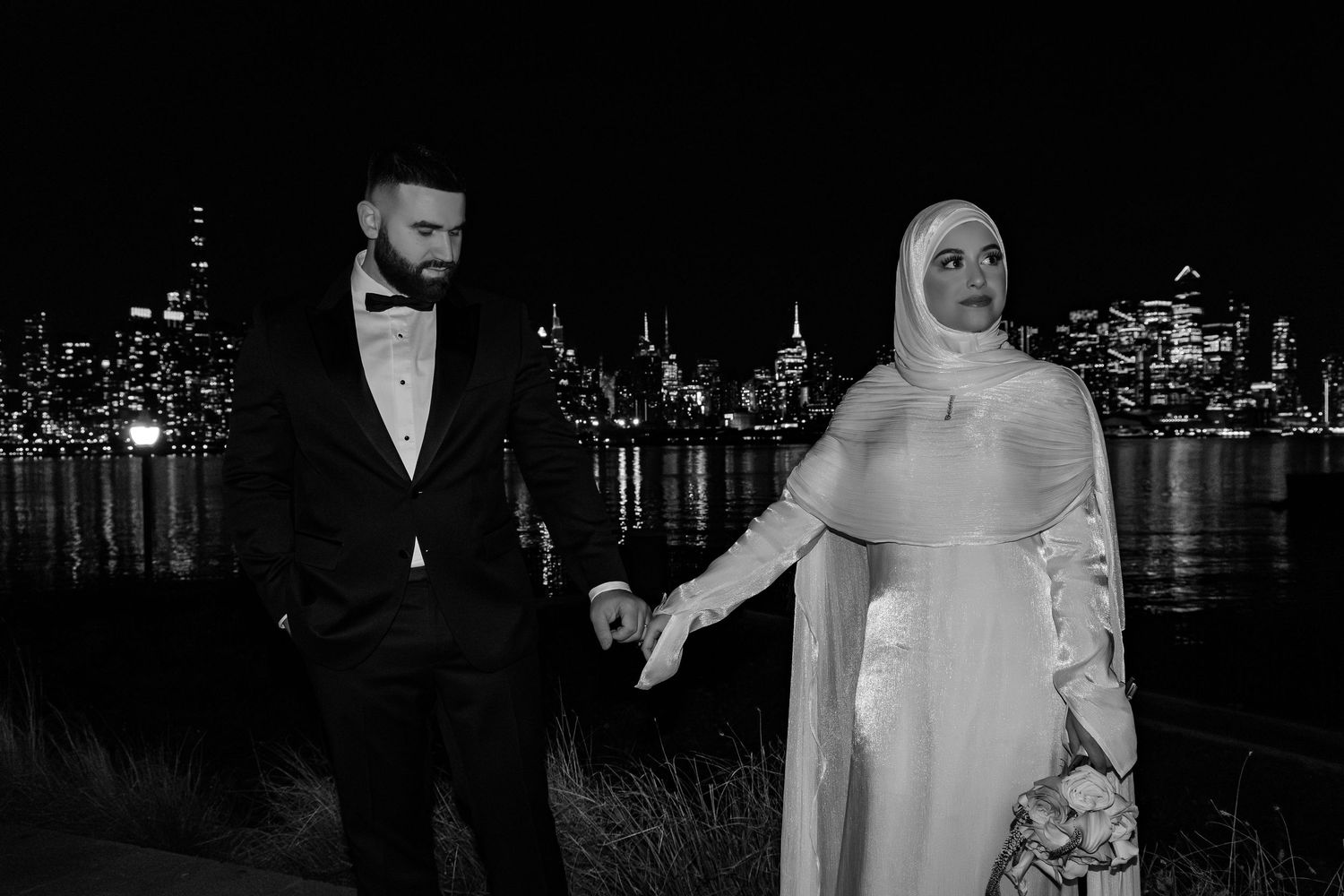 Black and white photo of Muslim bride and groom holding hands with city skyline at night.