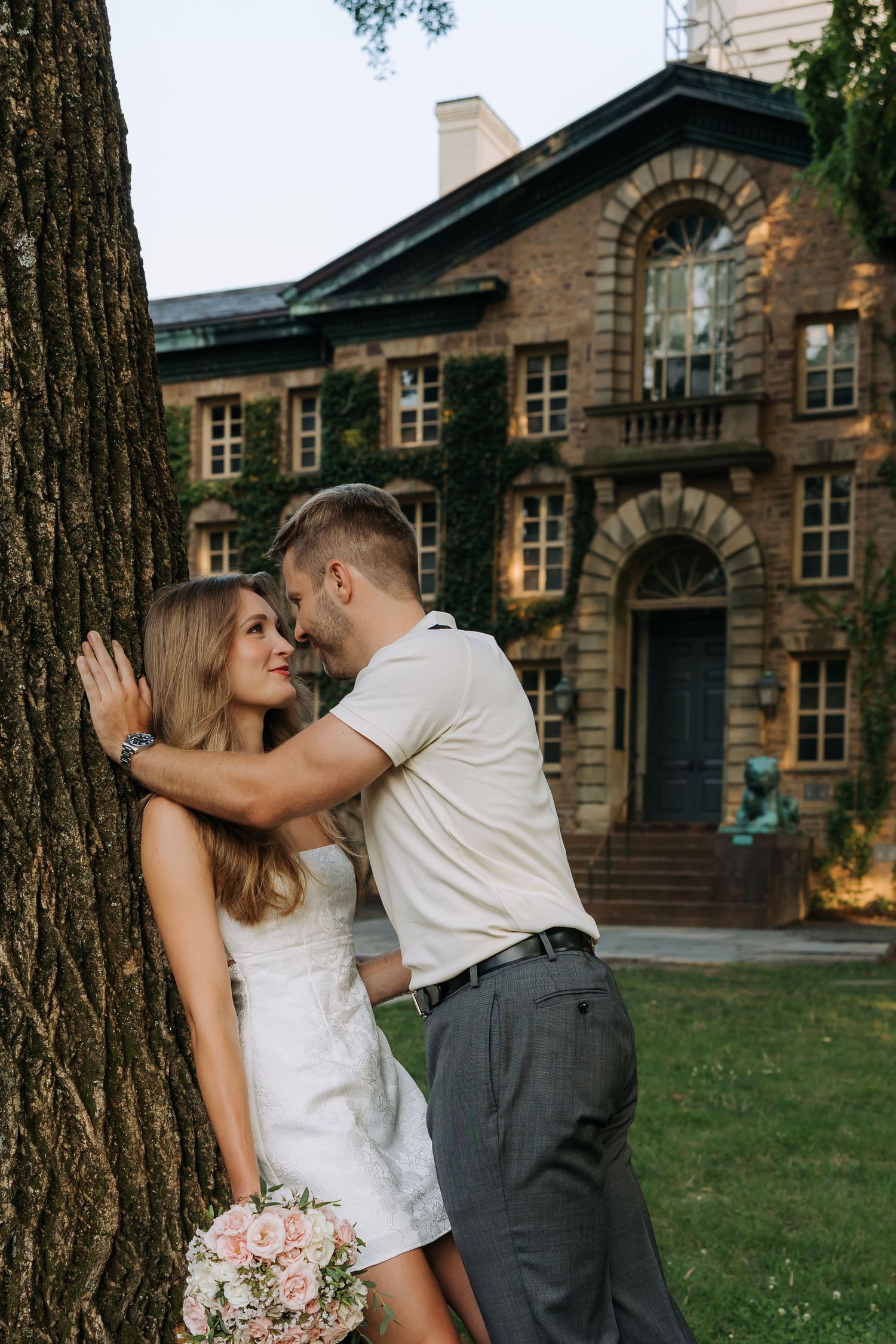 Couple sharing a romantic moment by a large tree, woman in white dress holding flowers, brick mansion in background.