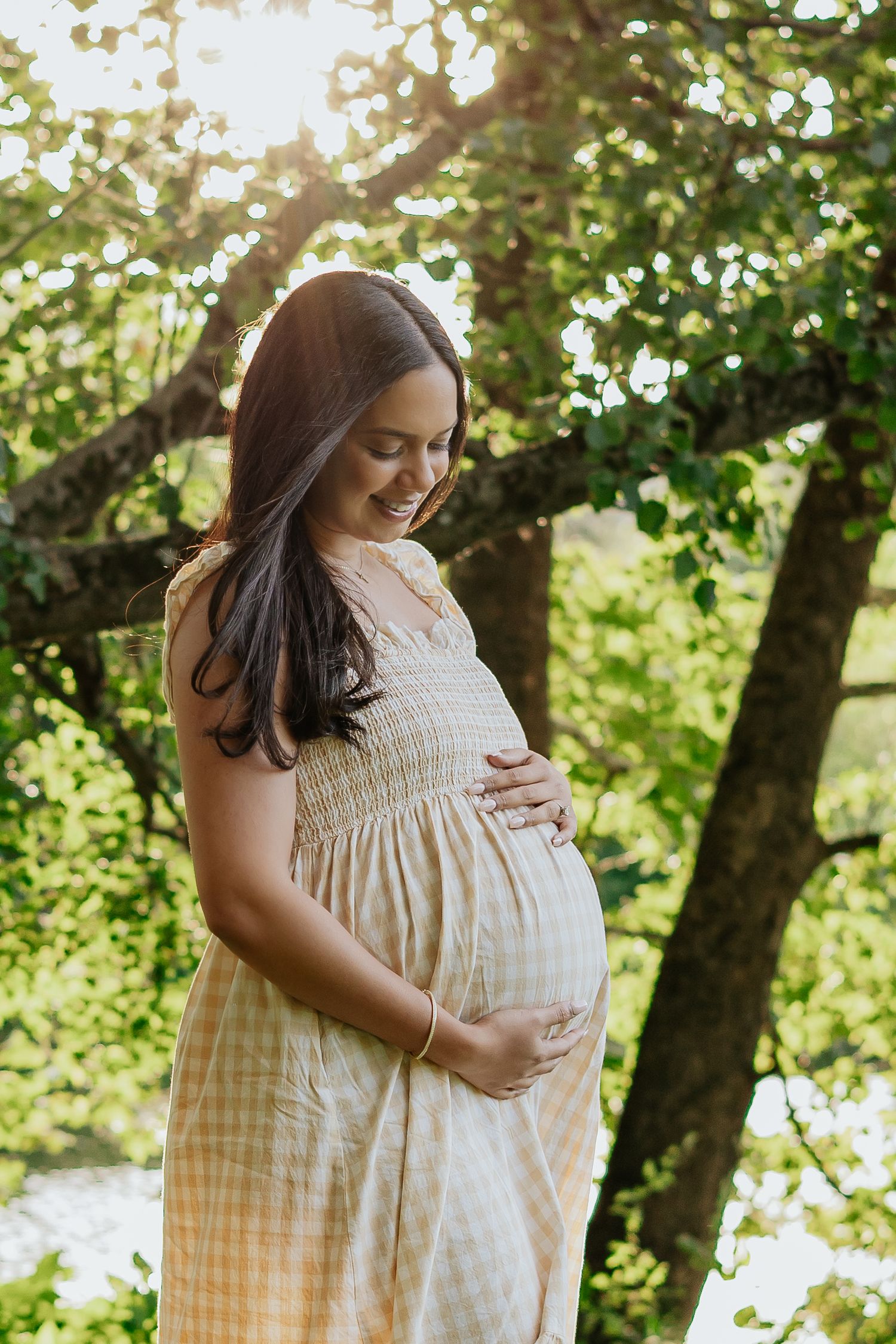 Smiling pregnant woman in yellow gingham dress tenderly holding her baby bump surrounded by sunlit green trees.