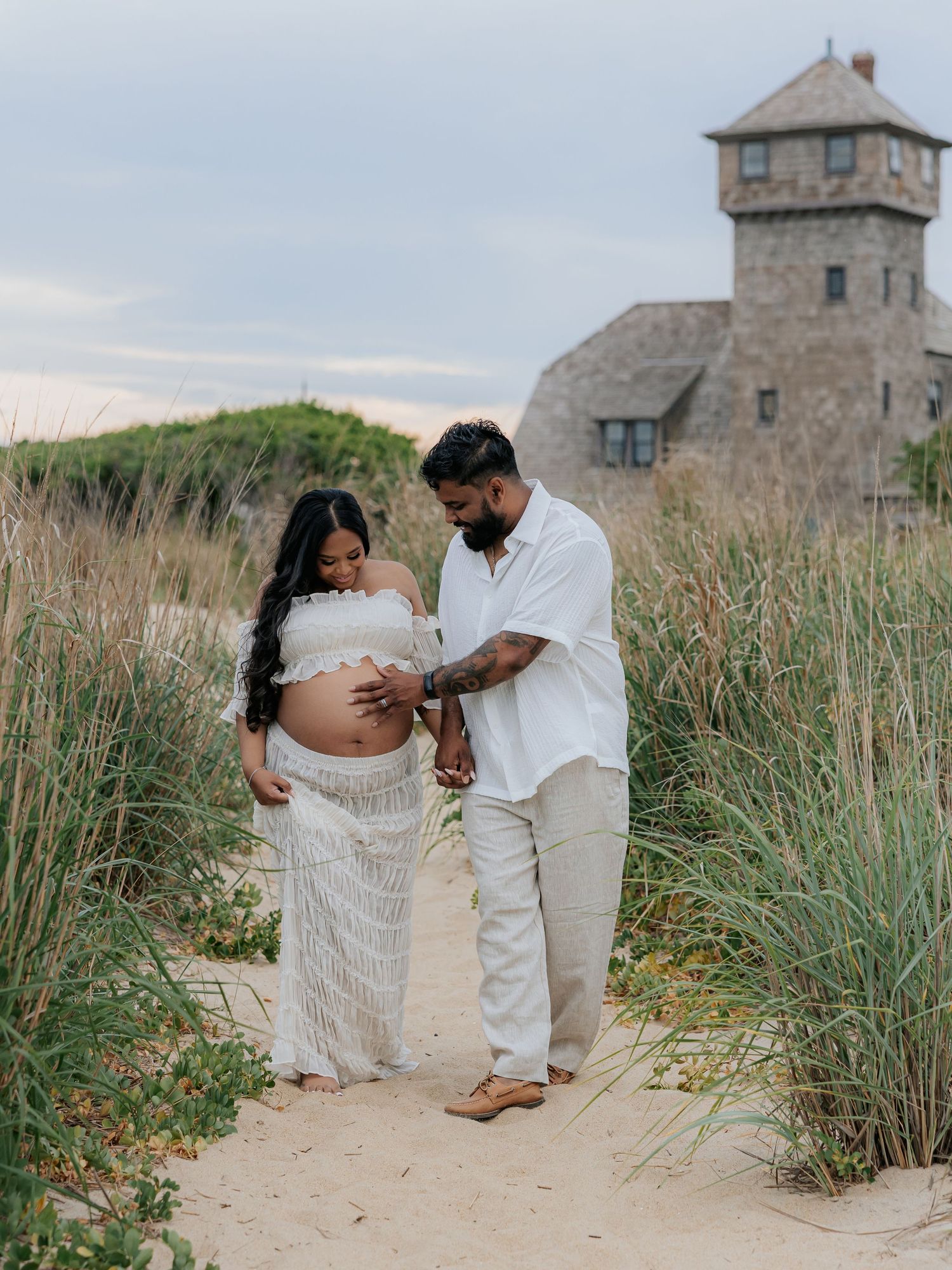 Pregnant couple in white outfits standing on sandy beach path surrounded by tall grass with a stone tower building in background.