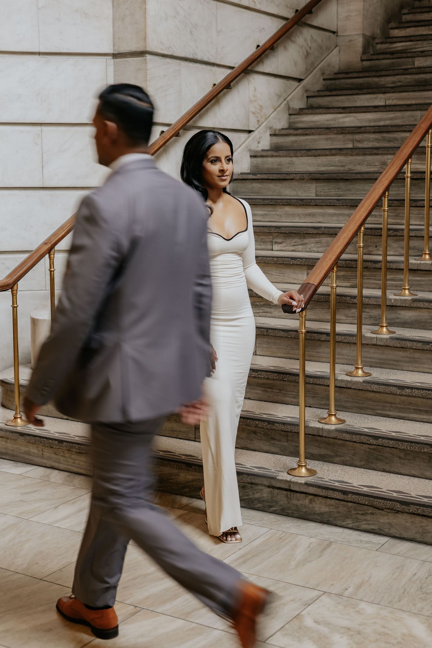 Bride in white gown holds groom's hand on marble staircase with gold railings.