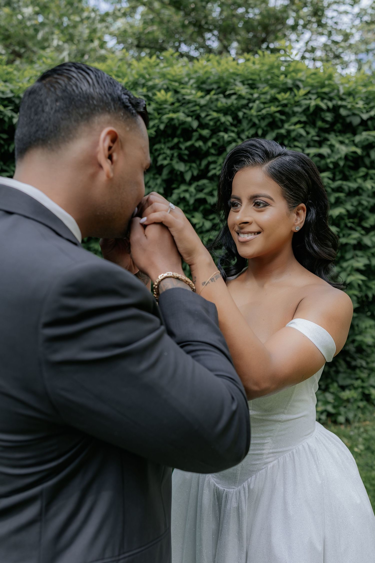 Groom kissing bride's hand while she smiles lovingly during outdoor engagement session in garden.