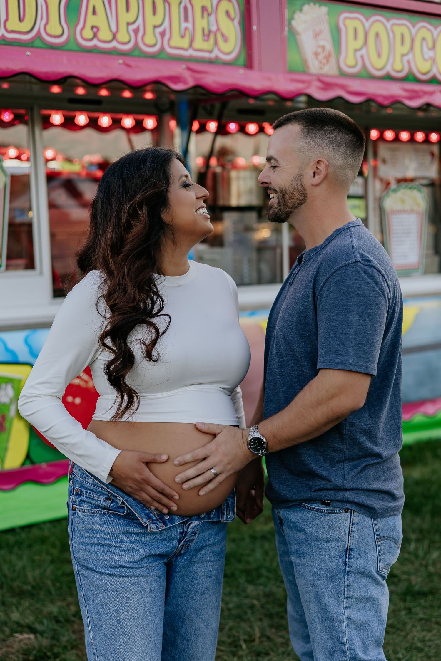 Smiling expecting couple at carnival, man tenderly holding pregnant partners baby bump in front of food stand.