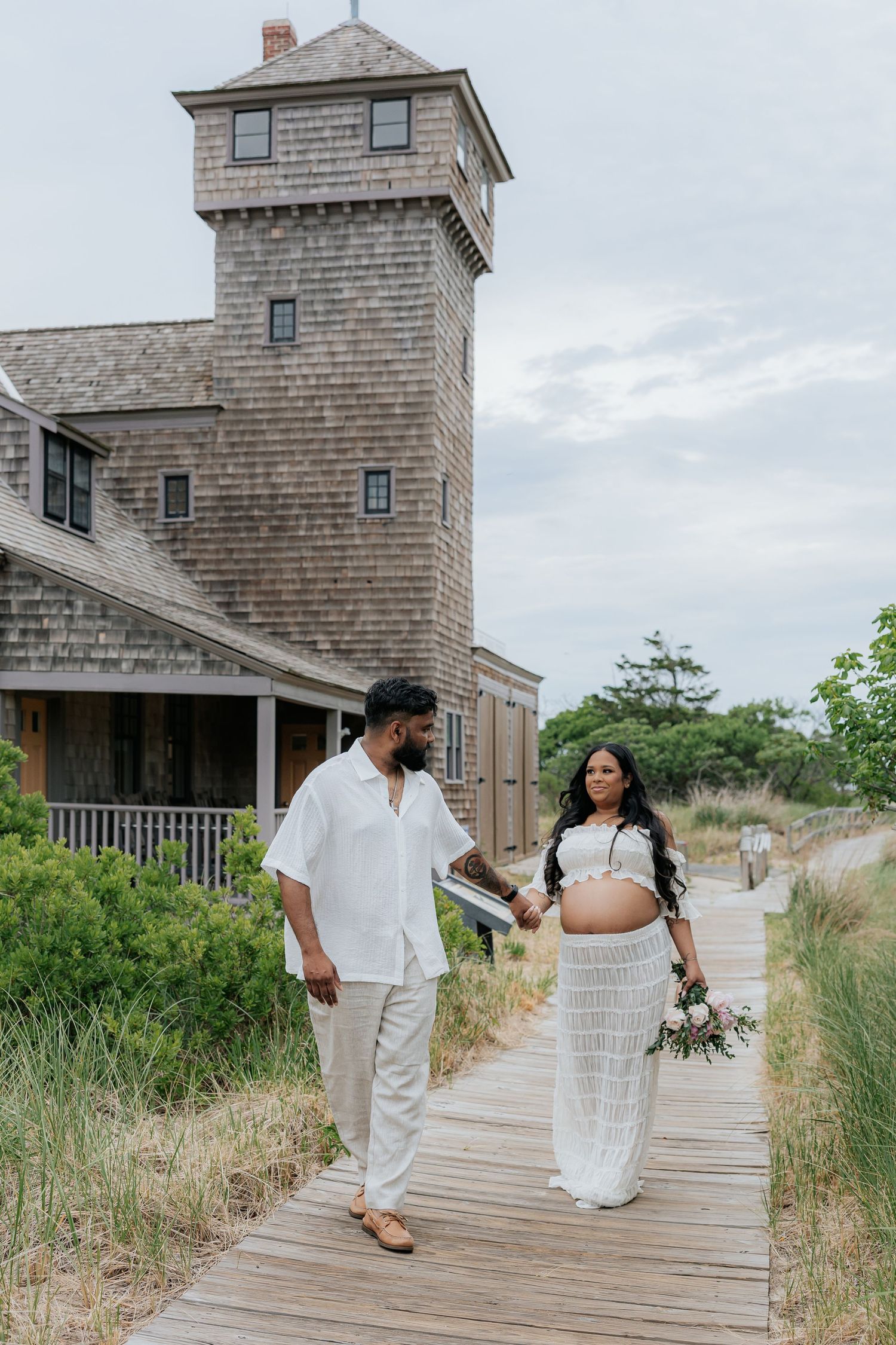 Pregnant couple in white outfits walking on a boardwalk near a rustic shingle-style building on a cloudy day.